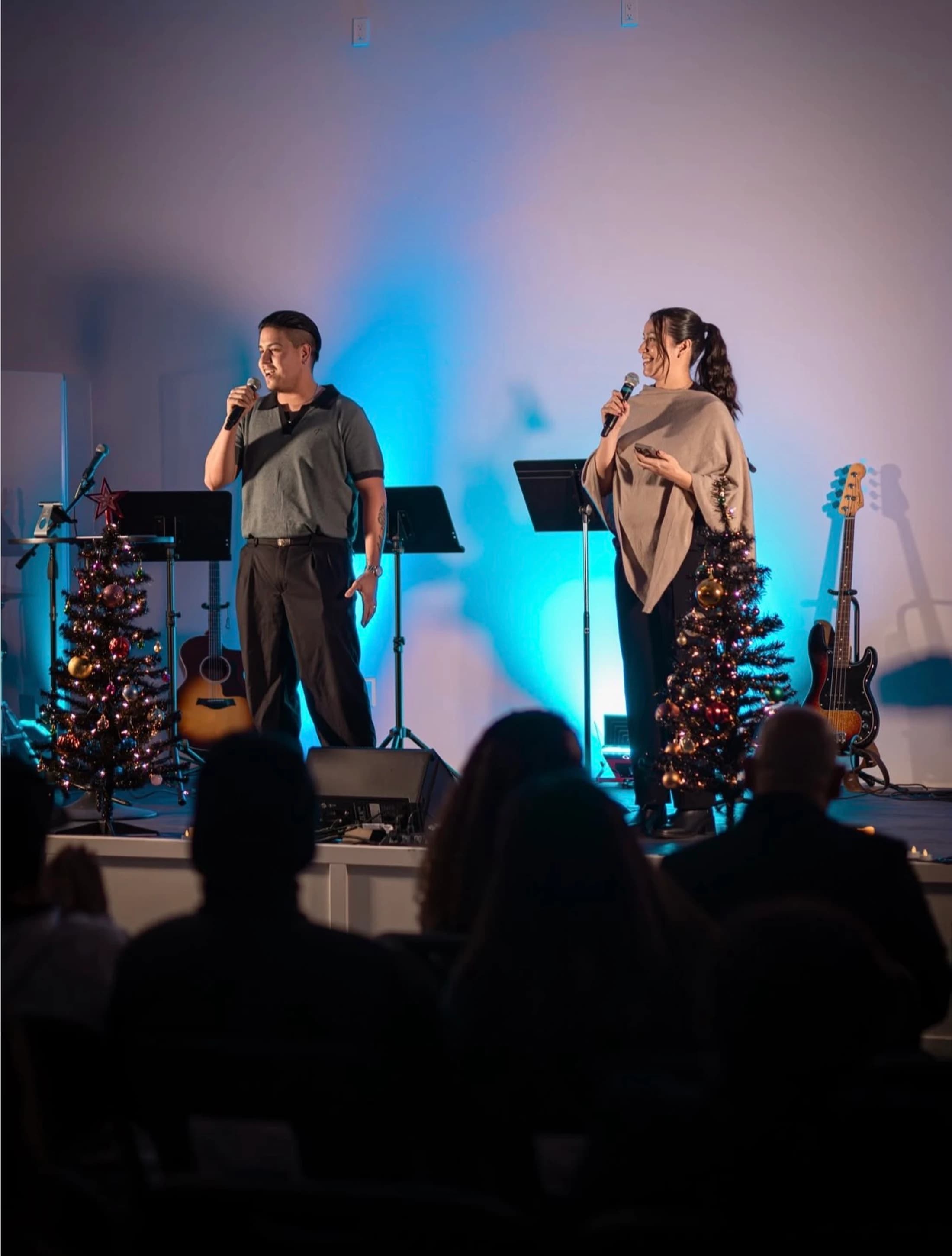A man and a woman stand on a stage with blue lighting, speaking into microphones. The stage is decorated with small Christmas trees for a holiday event.