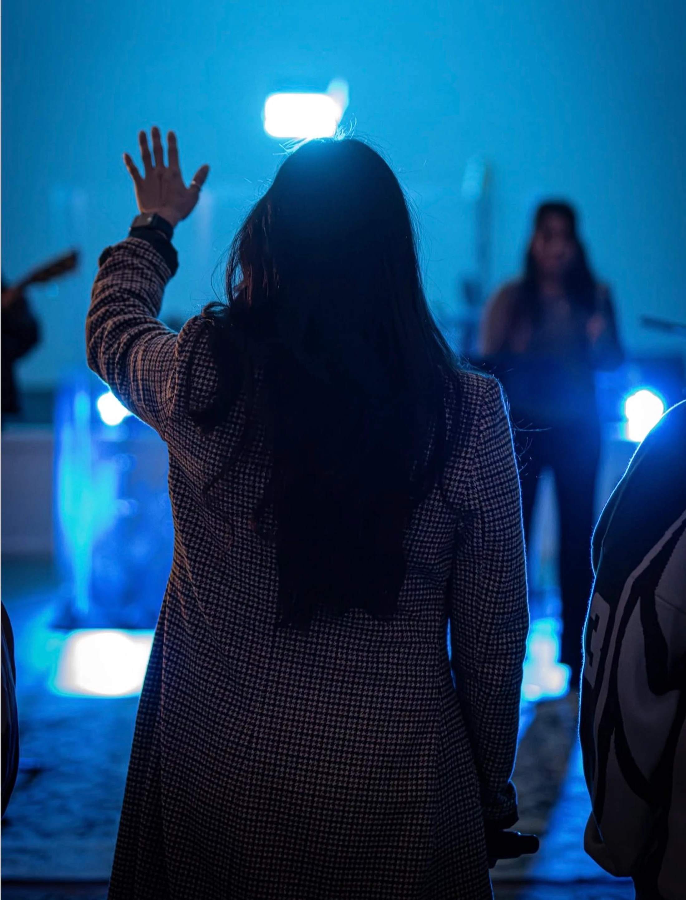 A rear view of a woman with long dark hair in a patterned coat, raising her hand during a worship service or concert illuminated by bright blue lights.