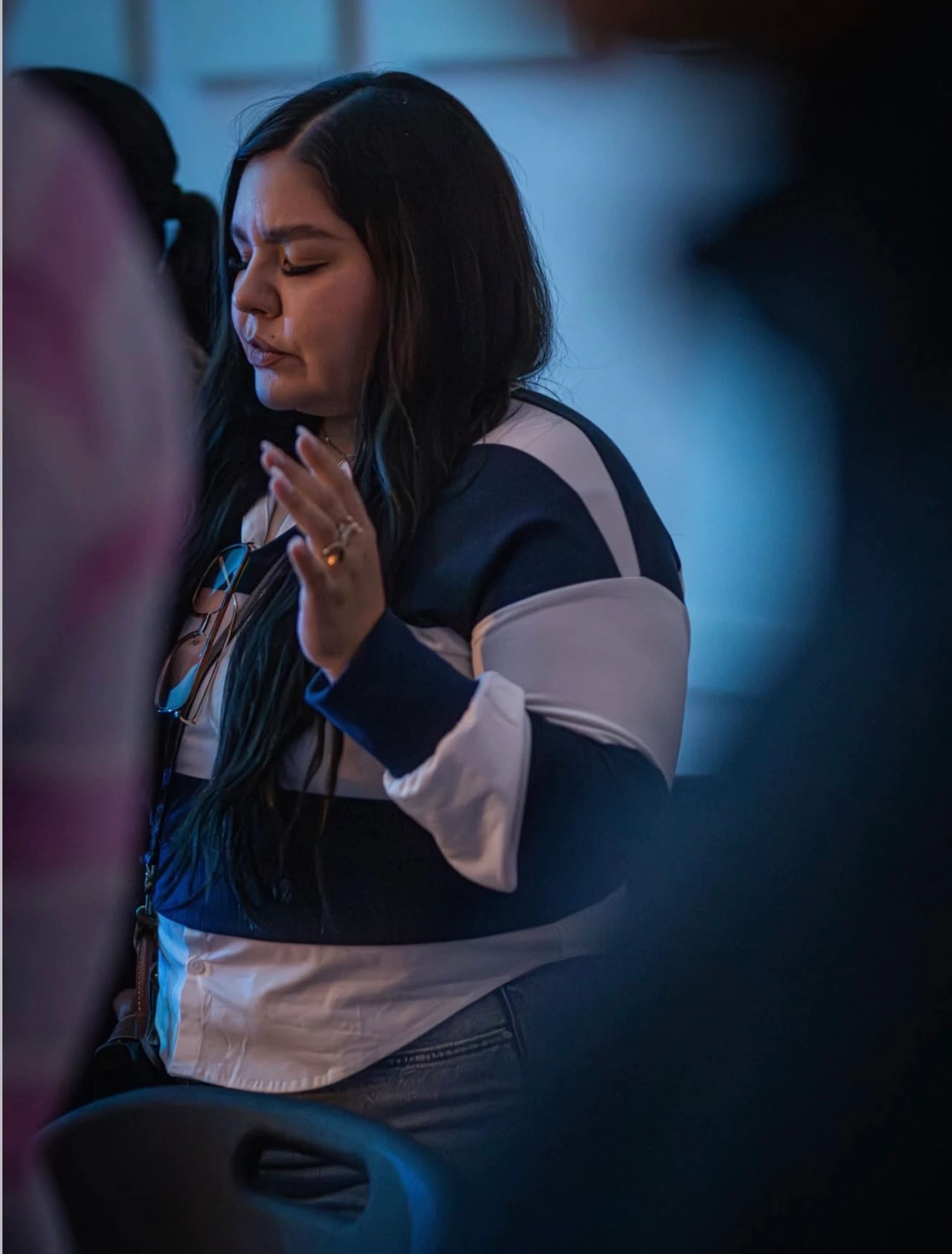A woman with dark hair stands with her eyes closed and a hand raised in worship. She is in a dimly lit room with blue lighting.