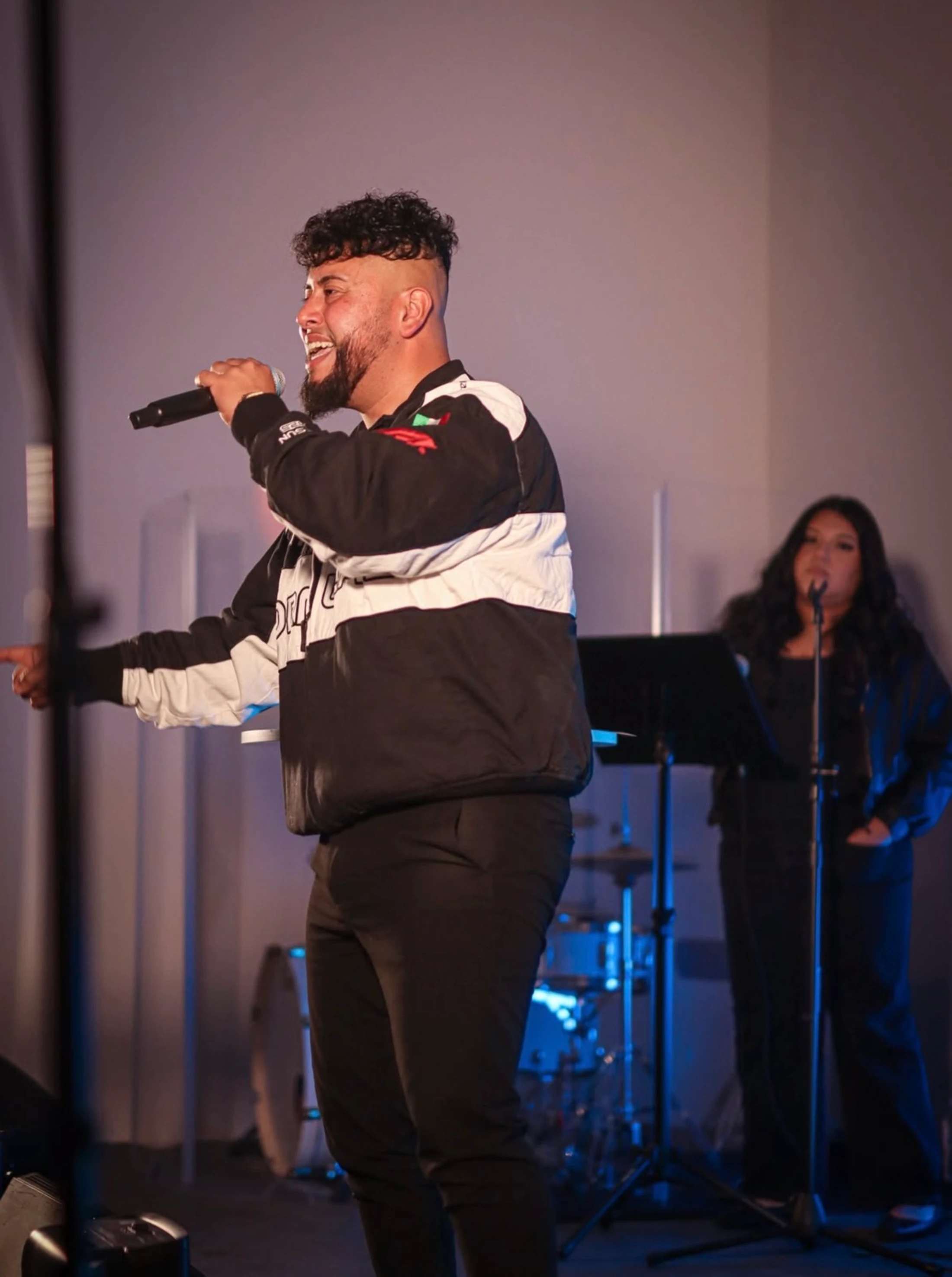 A male singer with a beard and curly hair smiles while singing into a microphone on stage, with a female backup singer and drum set in the background.