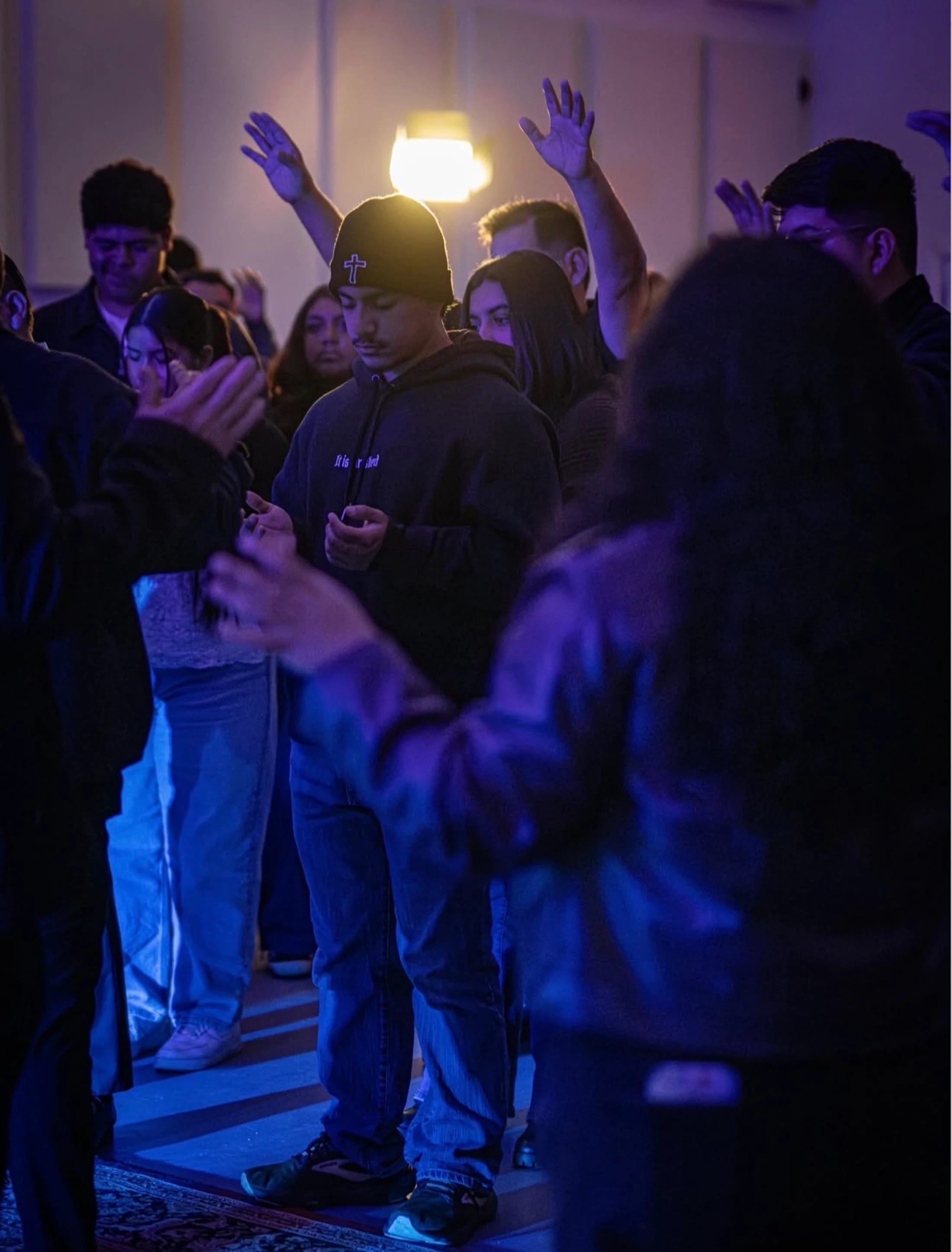 A diverse group of people worshiping in a dimly lit room with blue light. A young man in a beanie with a cross is in focus, praying with his hands clasped.