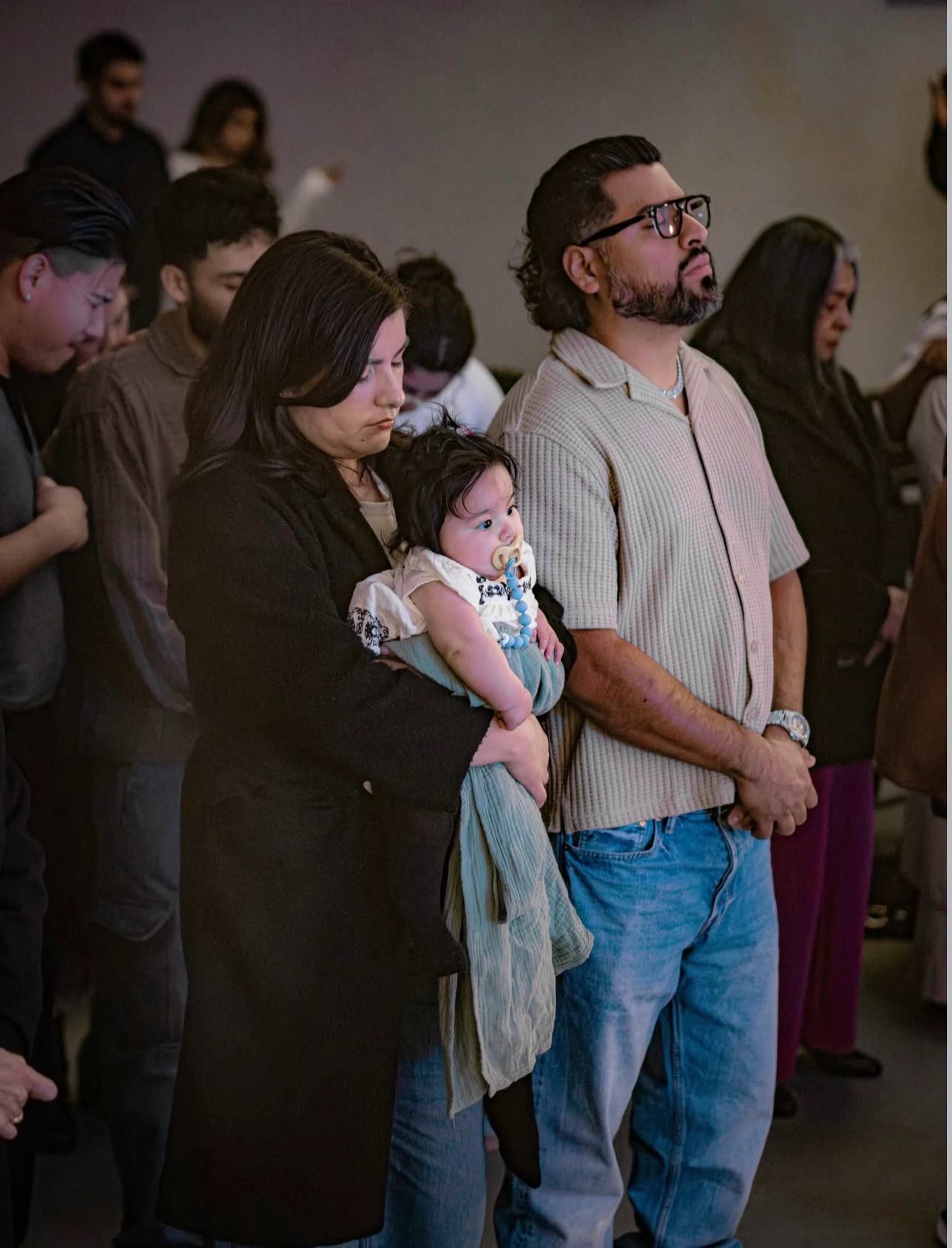 A family stands in a crowd during a gathering. A woman in a black coat holds a baby with a pacifier while a man in glasses looks forward thoughtfully.