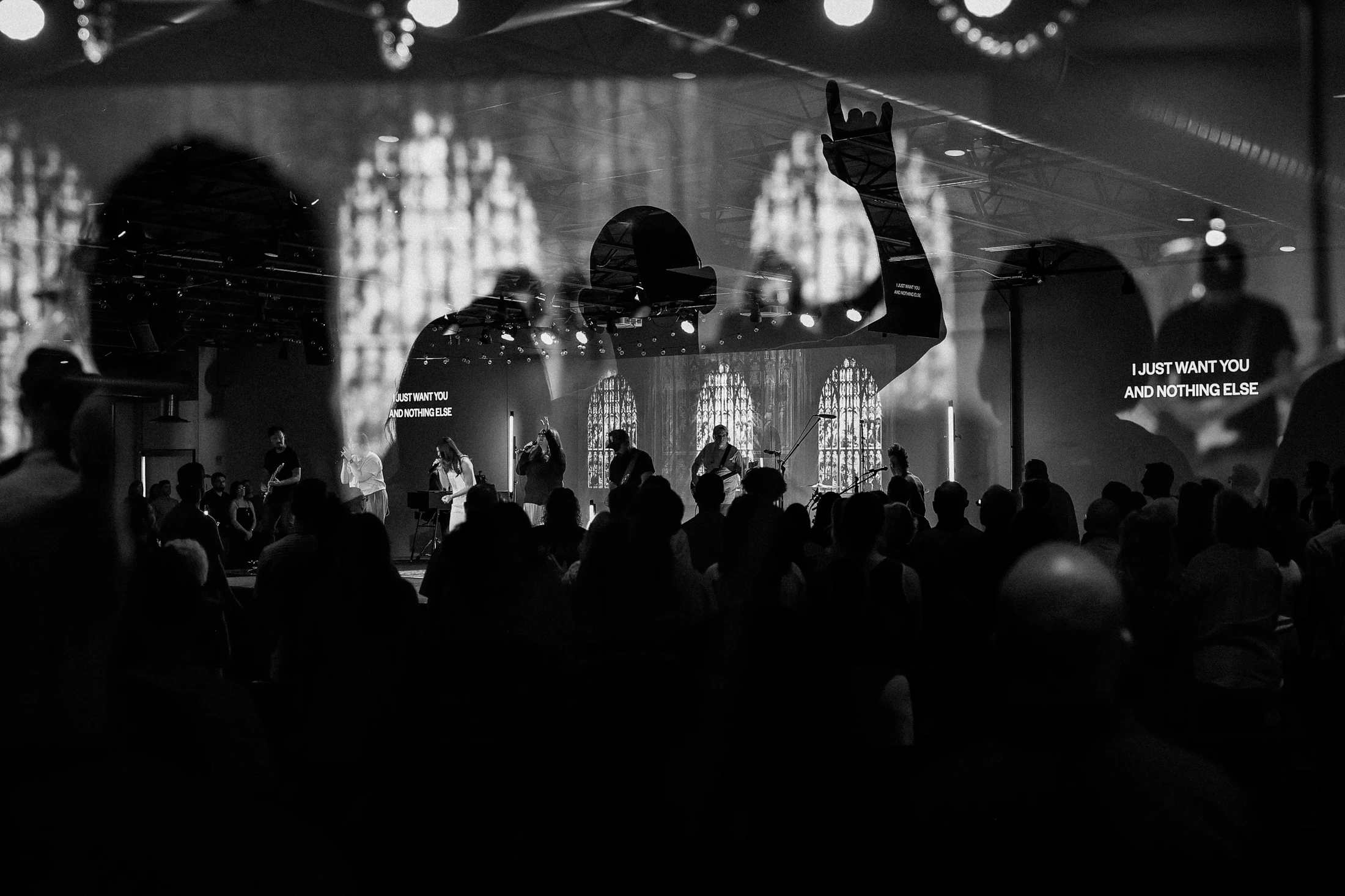 Black and white photo of a worship service. An audience watches a band on stage, with a large double exposure of a silhouetted figure with a raised hand.
