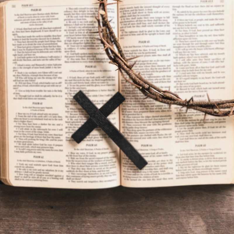 An open Bible lies on a wooden surface, with a crown of thorns and a small black cross resting on its pages. The photo is taken from directly above.