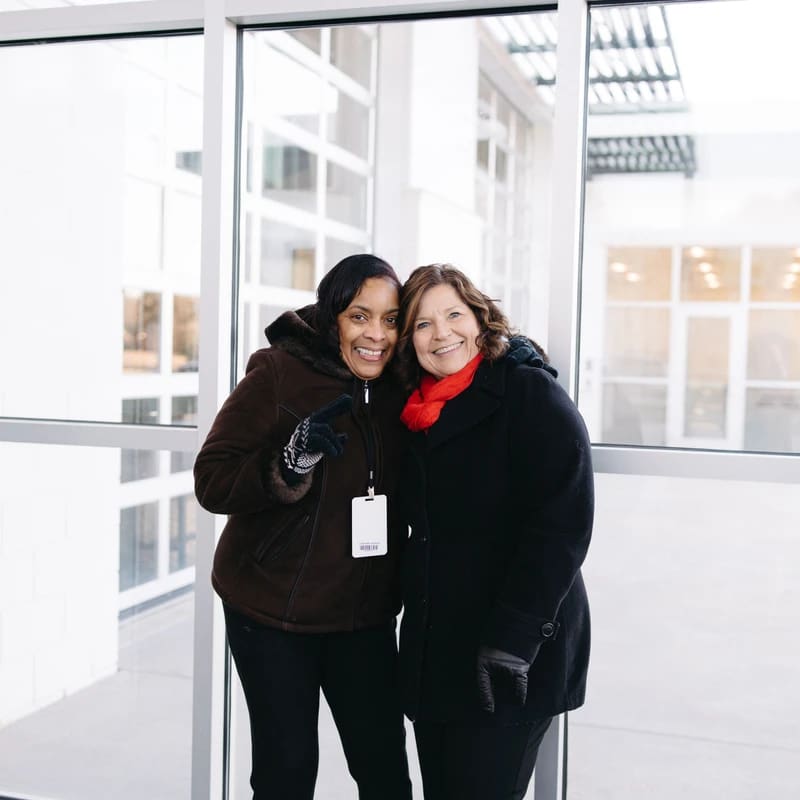 Two women, one Black and one white, stand smiling in front of a large window. They are wearing winter coats, gloves, and a red scarf.