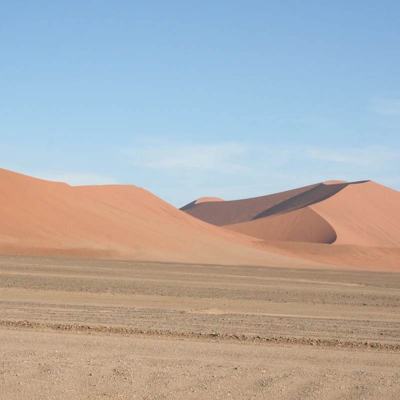 Large, reddish-orange sand dunes rise from a flat, sandy plain under a clear blue sky, creating a vast and arid desert landscape.