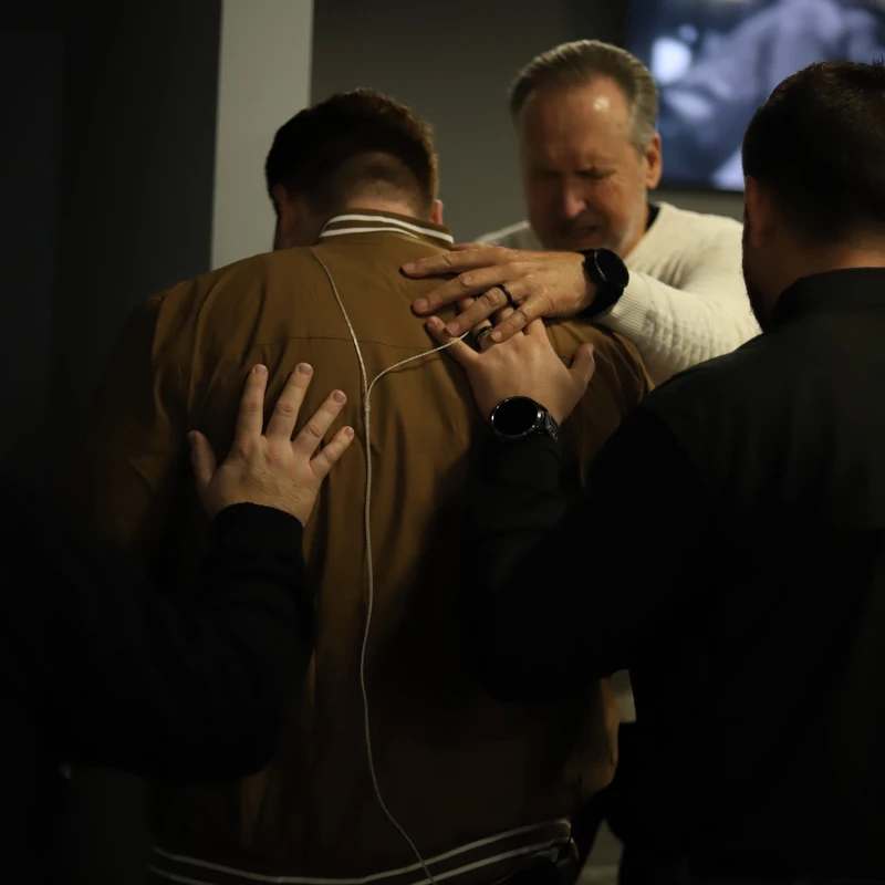 Several people place their hands on the back of a man in a brown jacket in a supportive, prayerful gesture. An older man with his eyes closed is among them.