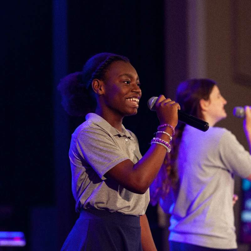 A young Black girl with a smiling face sings into a microphone on stage, with another girl singing in the background under purple and blue lights.