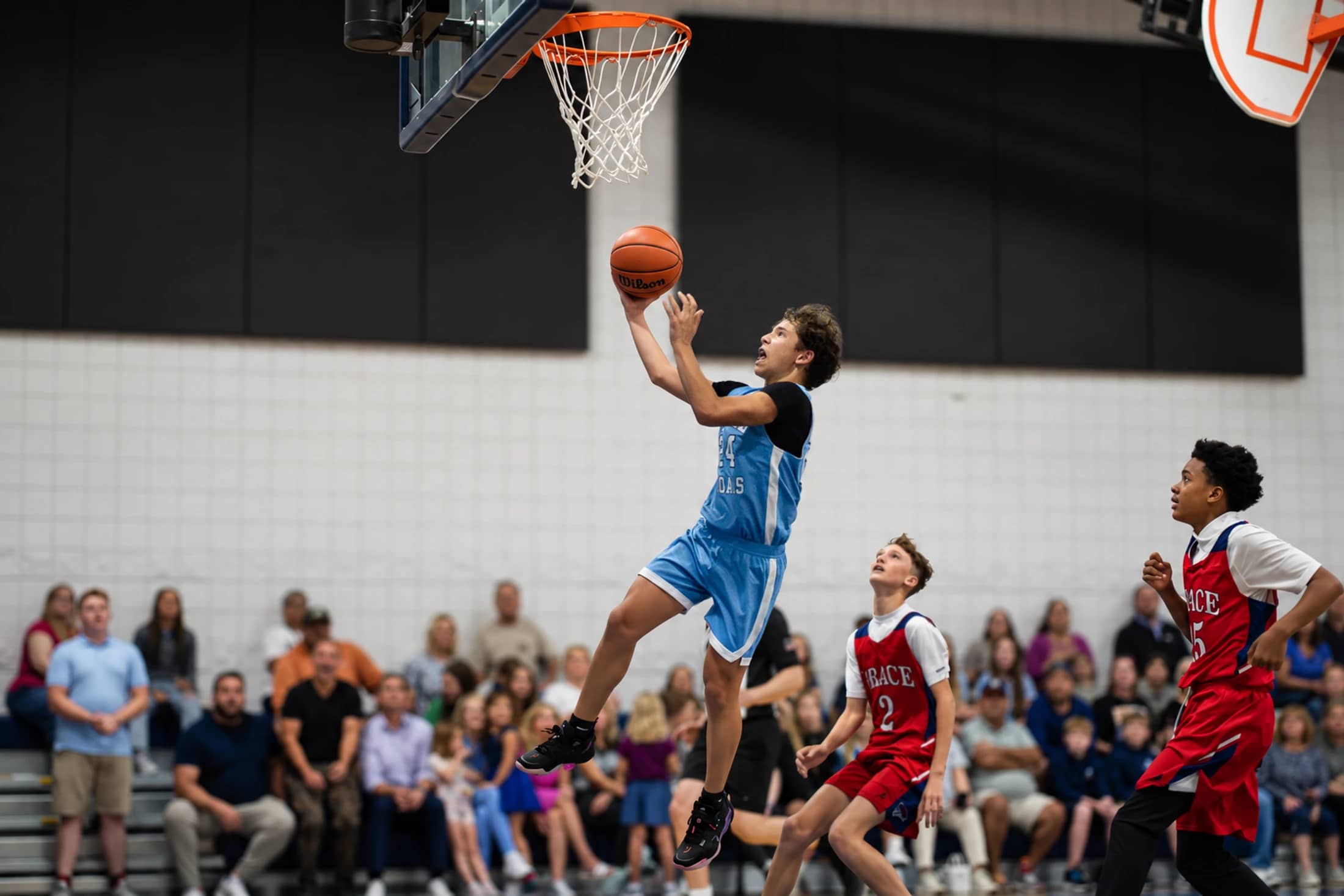 A young basketball player in a light blue uniform jumps towards the hoop to make a layup during a game as two opposing players in red watch.