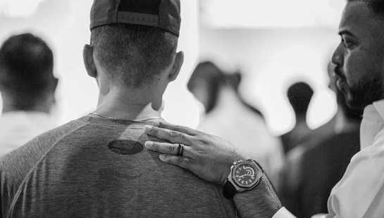 A black and white close-up photo of a man's hand resting on another man's shoulder in a supportive gesture. The man being comforted is seen from behind.