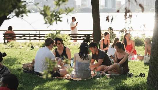 A group of young friends sit together on a picnic blanket in a grassy park by the water, enjoying a sunny afternoon with the city skyline in the background.