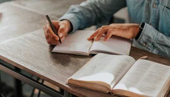 A close-up of a person in a blue shirt writing in a notebook at a wooden desk, with an open book, possibly a Bible, in the foreground.
