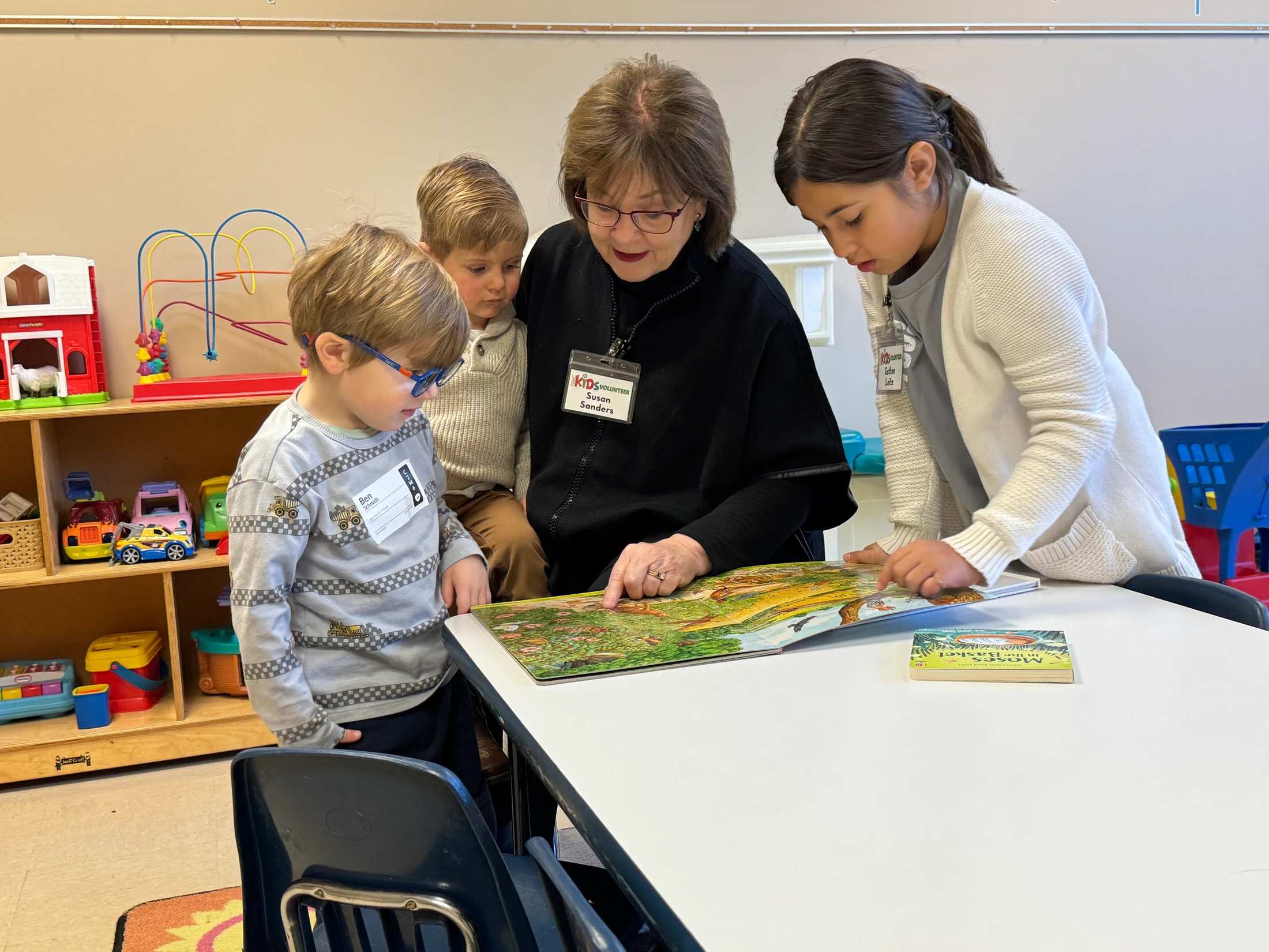 An adult volunteer and three children are gathered around a table in a classroom, looking at and pointing to pictures in a colorful, open book.