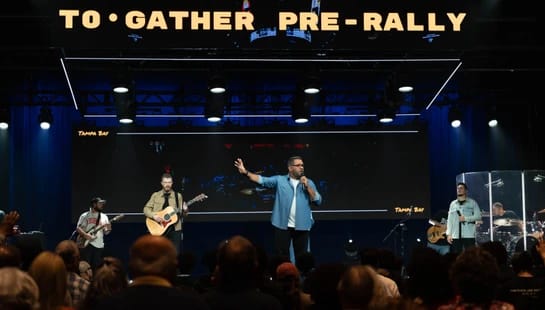 A man speaks on a stage with a band to a large audience. A screen behind them reads 'TO•GATHER PRE-RALLY' and 'Tampa Bay'.