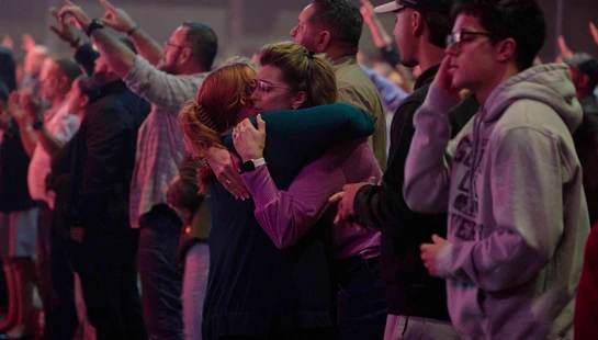 Two women share an emotional hug in the middle of a crowd at an event. Other people stand around them, some with their hands raised in an act of worship.