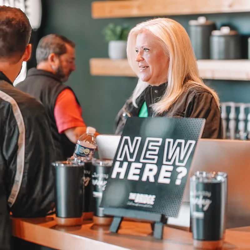 A smiling blonde woman welcomes a guest at an information desk. A sign on the counter reads 'NEW HERE?'.
