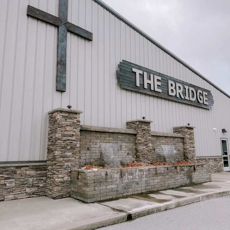 The exterior of a modern, light gray building with a sign that reads 'The Bridge'. A large wooden cross is on the wall above a stone water fountain.