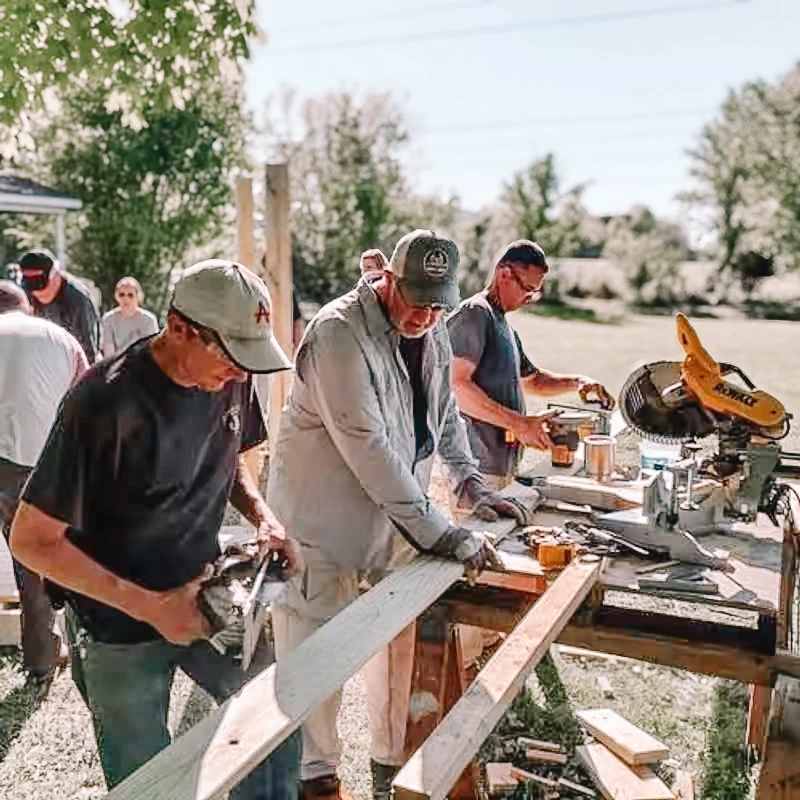 A group of men work together on a construction project outdoors on a sunny day. They use a miter saw and other tools to cut wood planks on a workbench.