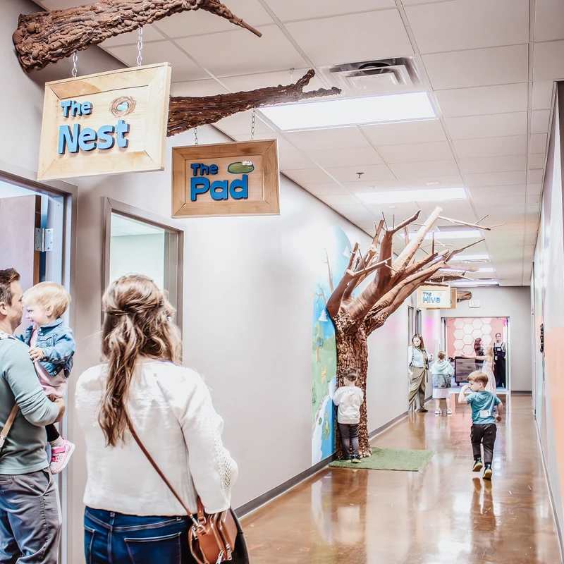A family stands in a brightly lit hallway of a children's center. The hall has nature-themed decor, including signs for 'The Nest' and 'The Pad'.