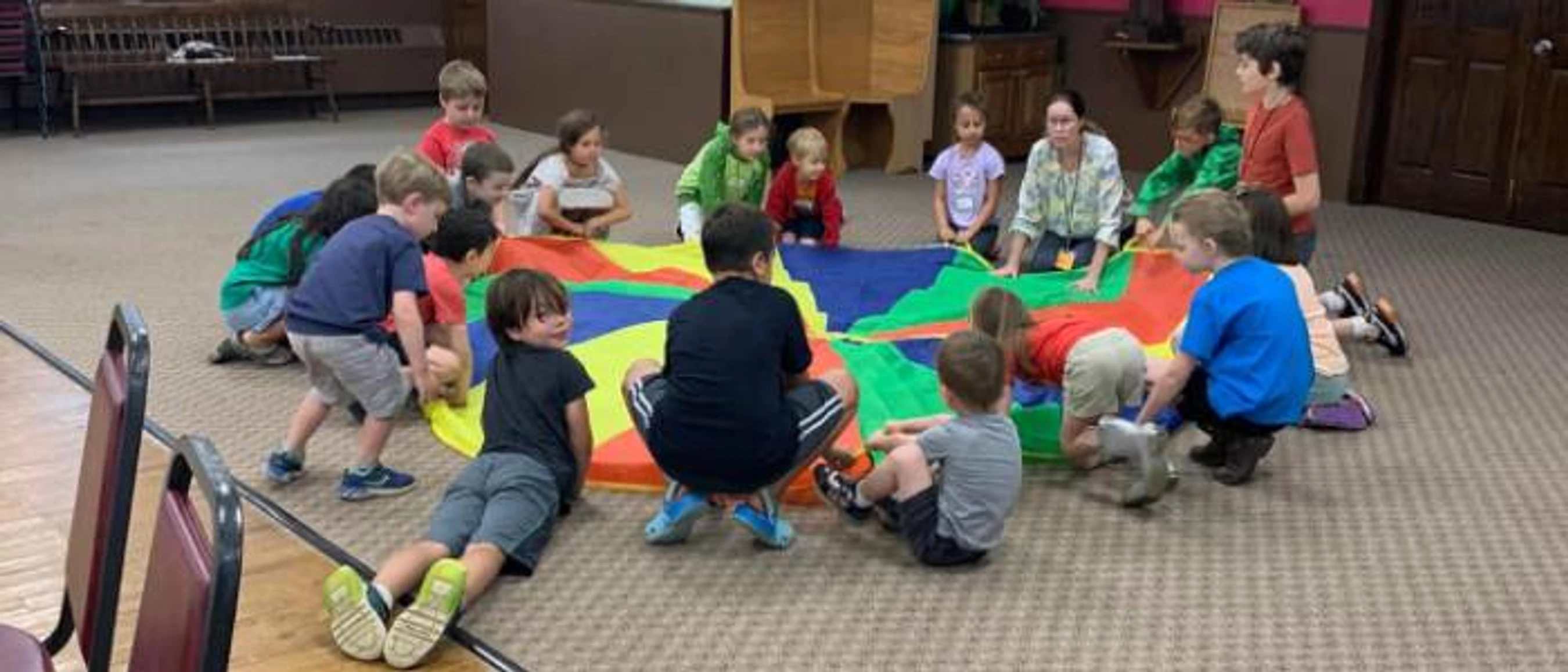 A group of young children and an adult sit in a circle on a carpeted floor, holding the edges of a large, colorful play parachute during an activity.