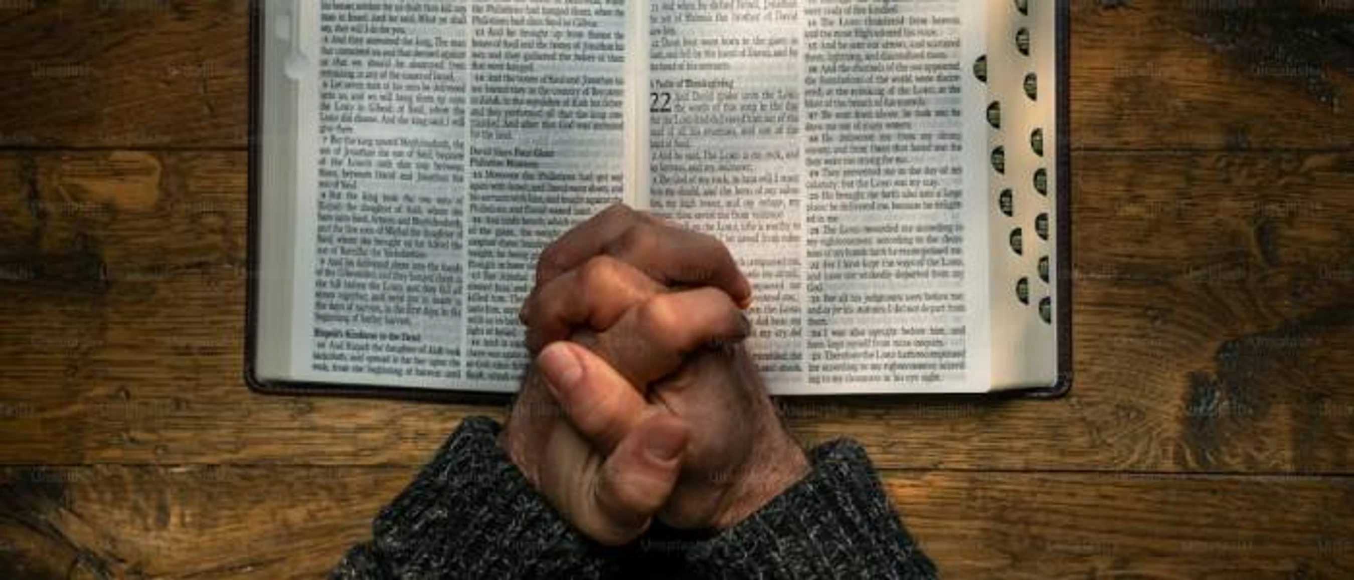An overhead view of a person's hands clasped in prayer, resting on an open Bible which is on a dark wooden table.