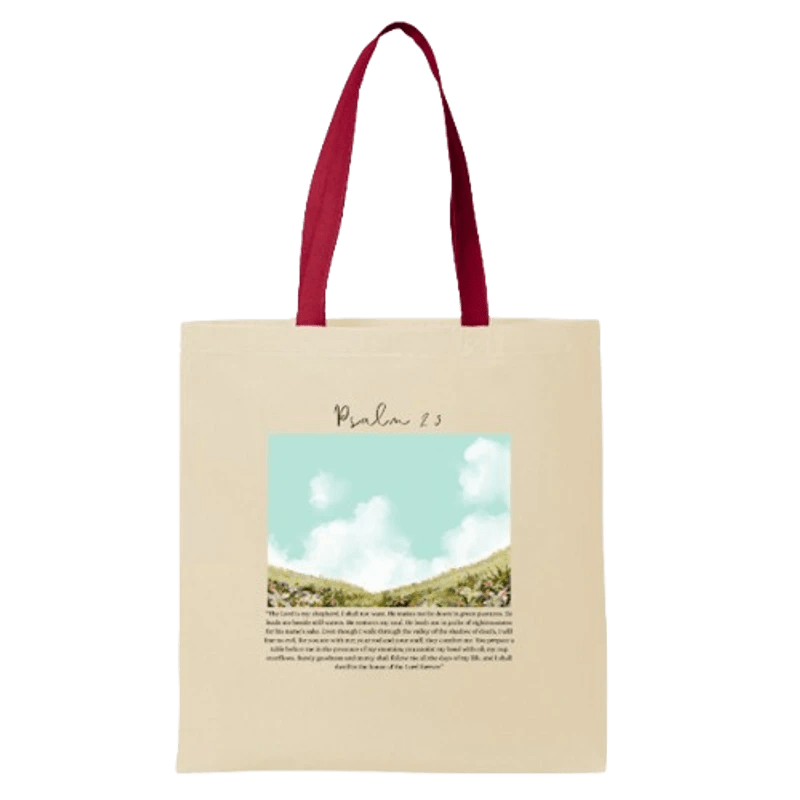 A cream-colored canvas tote bag with red handles, featuring a printed design of Psalm 23 with a peaceful landscape of hills and a cloudy sky.