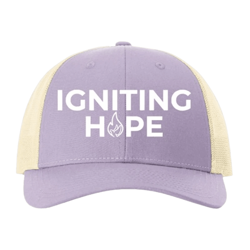 A lavender trucker hat with a cream mesh back. It features the text 'IGNITING HOPE' in white, with a flame icon replacing the letter 'O' in hope.