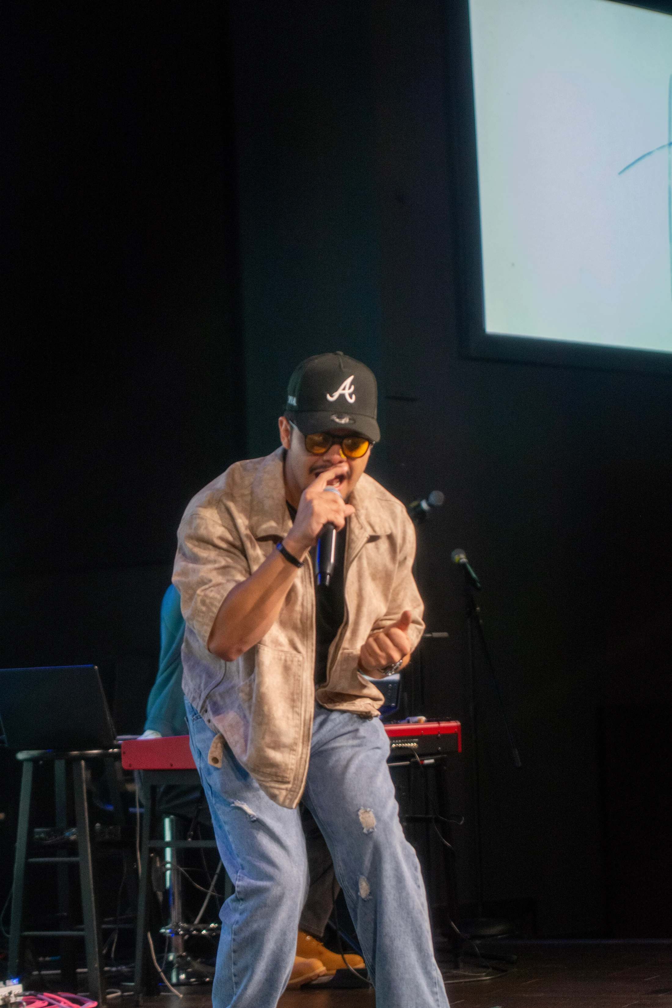 A man wearing a baseball cap and sunglasses sings into a microphone on a dark stage. He is wearing a jacket and ripped jeans.