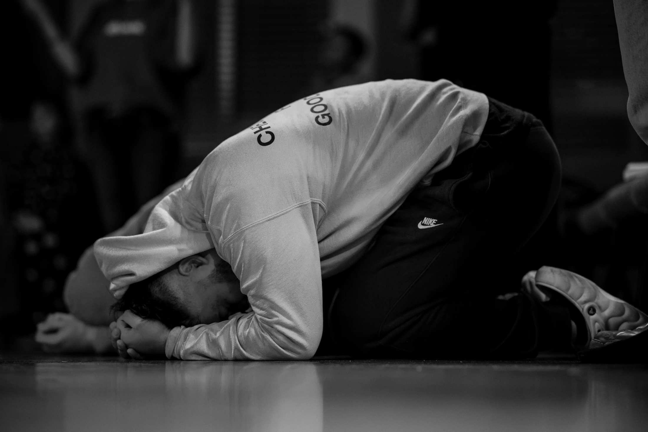 A black and white photo of a person in a hoodie kneeling on a reflective floor, curled over with their head bowed, suggesting exhaustion or contemplation.