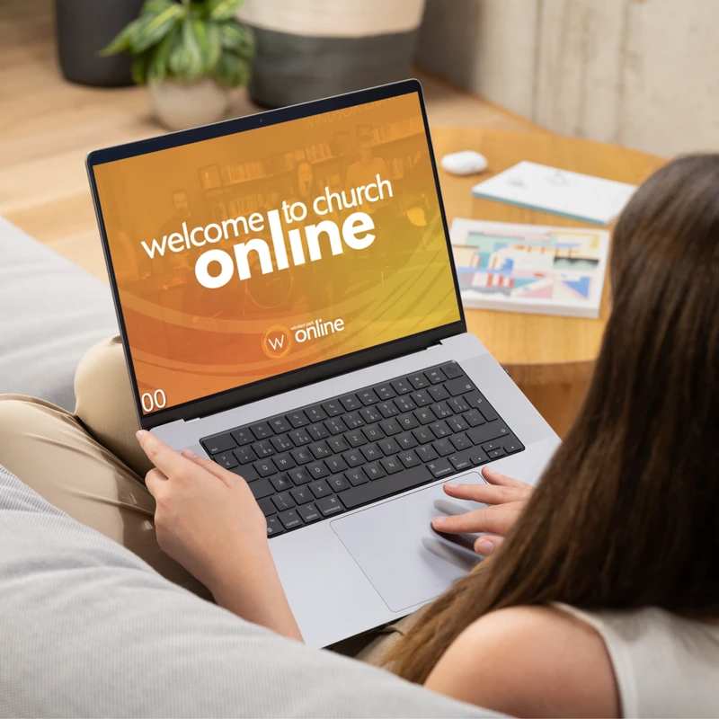 Over-the-shoulder view of a woman on a couch watching an online church service on her laptop. The screen says 'Welcome to church online'.