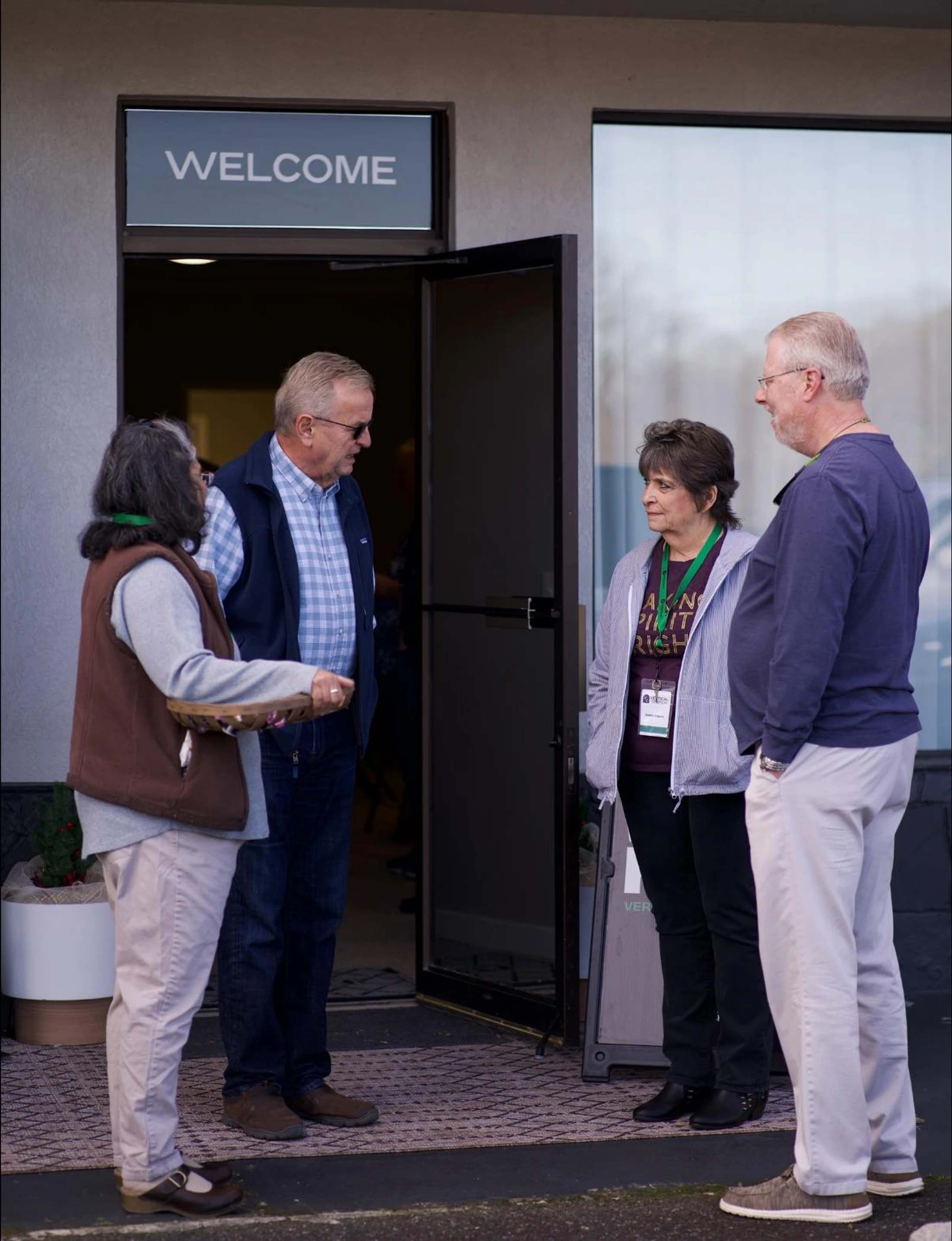 Four adults stand talking in pairs outside an open doorway with a 'WELCOME' sign above it. A woman on the left holds a small basket.