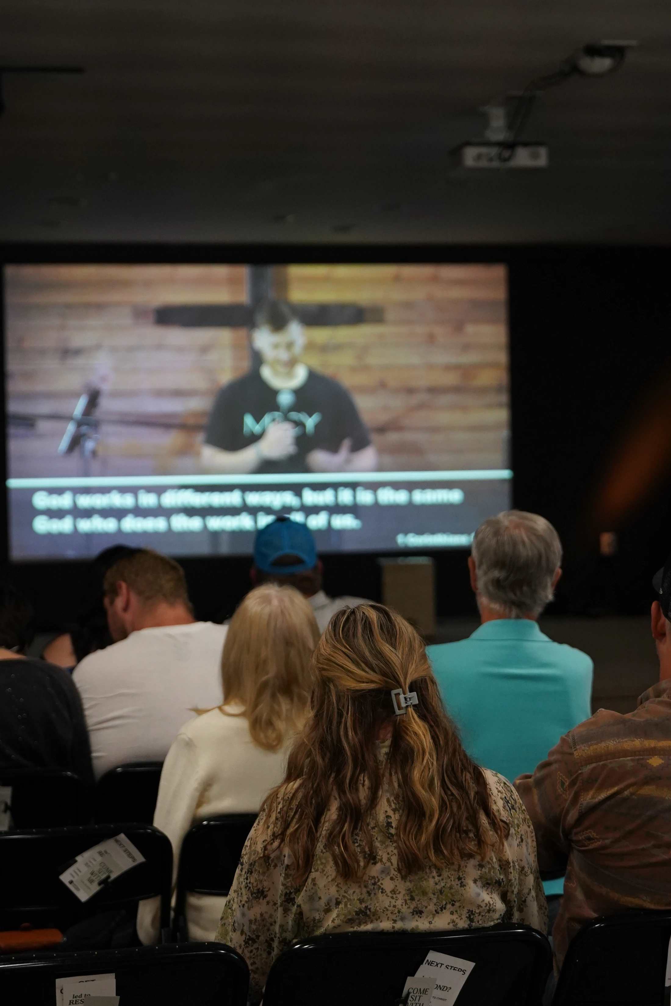 The backs of an audience sitting in chairs watching a man give a sermon on a large screen. The screen has subtitles and a large cross is visible.