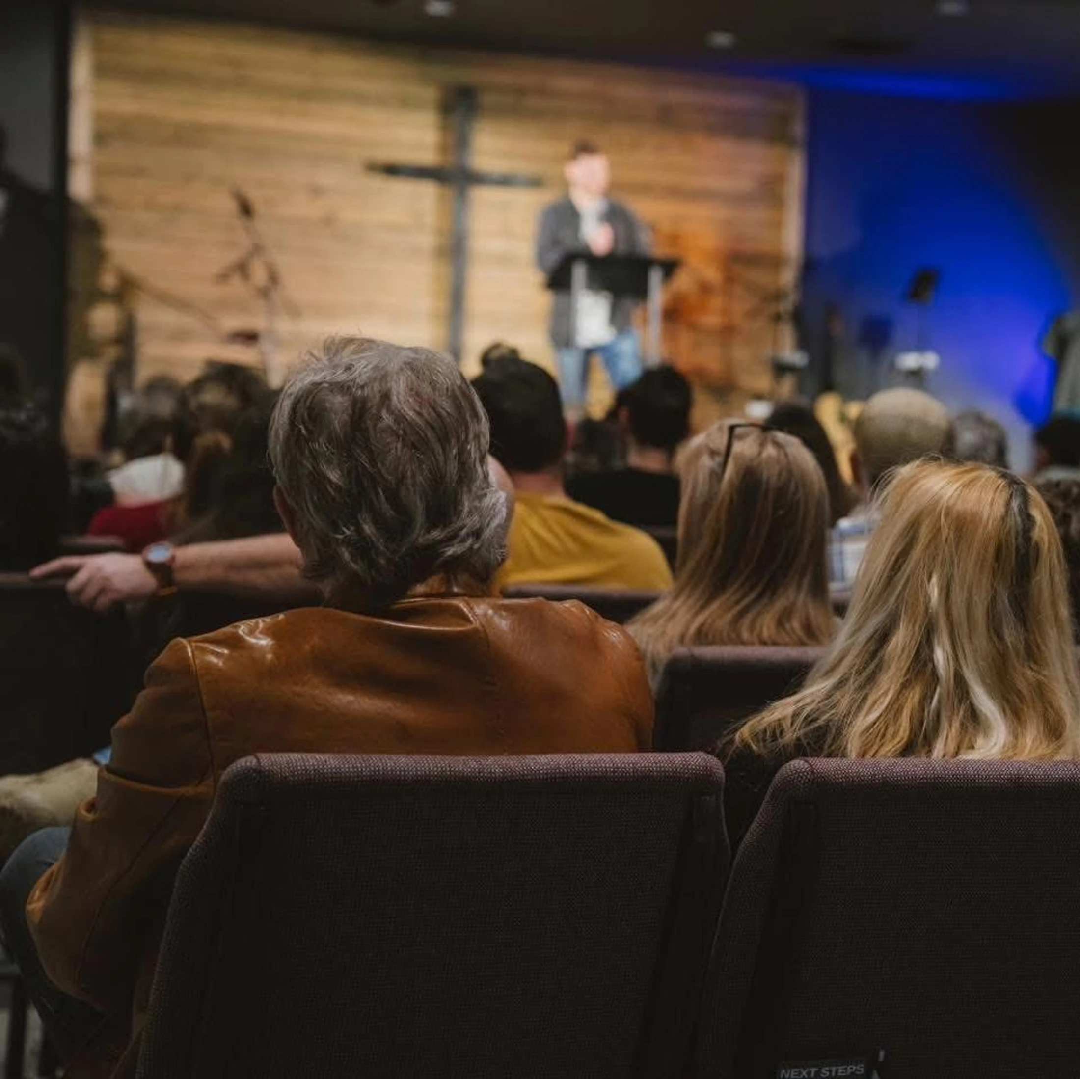 The view from behind a congregation sitting in chairs, watching a speaker on a stage with a large wooden cross on the wall behind him.
