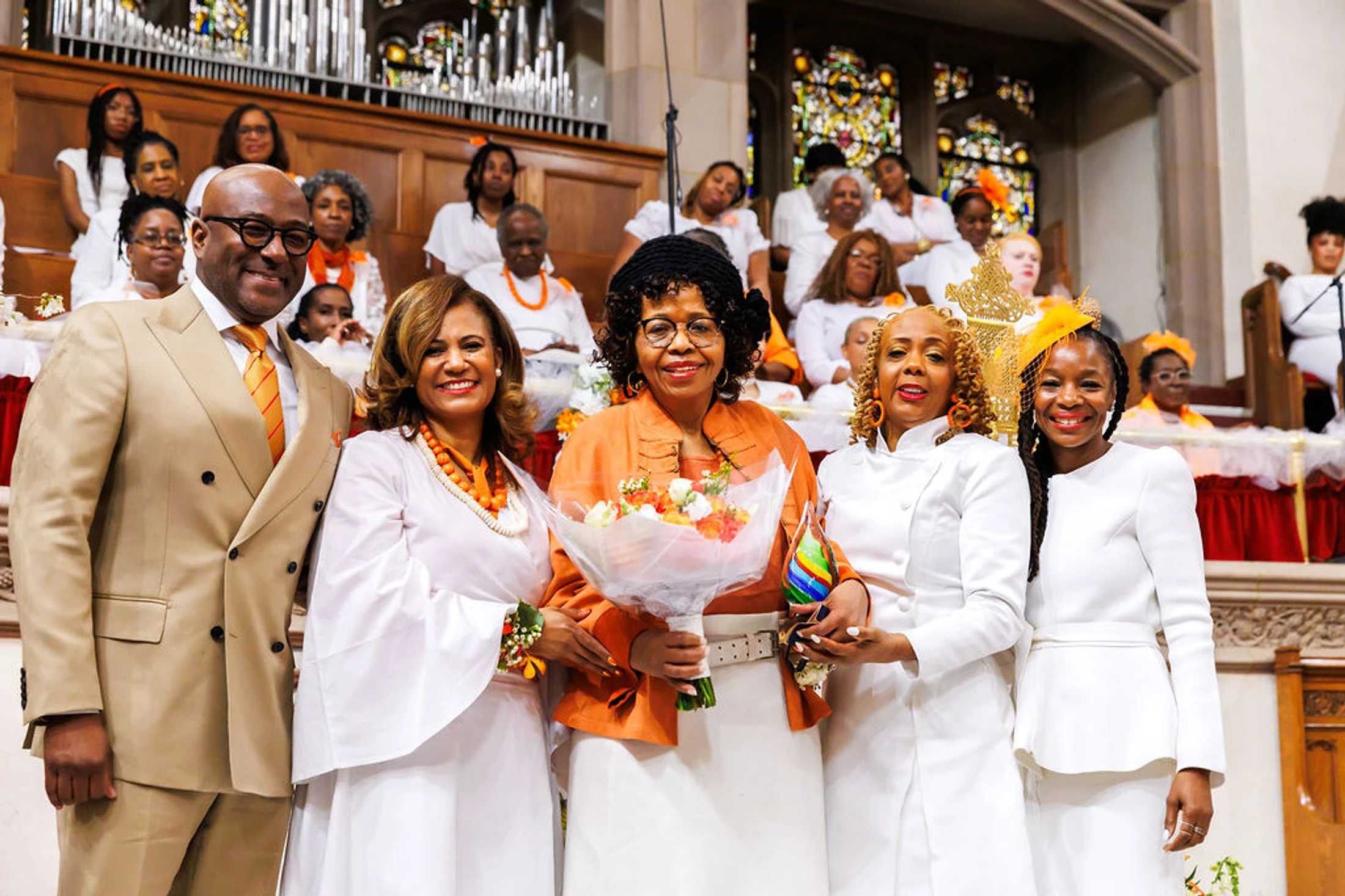 A group of five smiling Black adults pose for a photo in a church. A woman in the center holds flowers and an award, with a choir seated behind them.