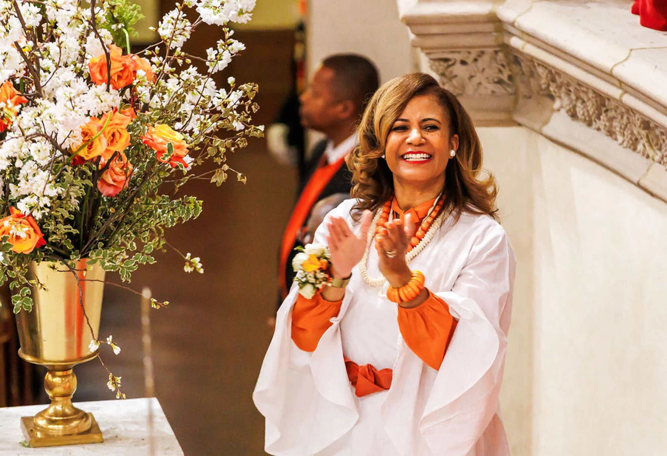 A woman with light brown hair, dressed in a white outfit with orange accents, smiles and claps her hands. Next to her is a floral arrangement in a gold vase.