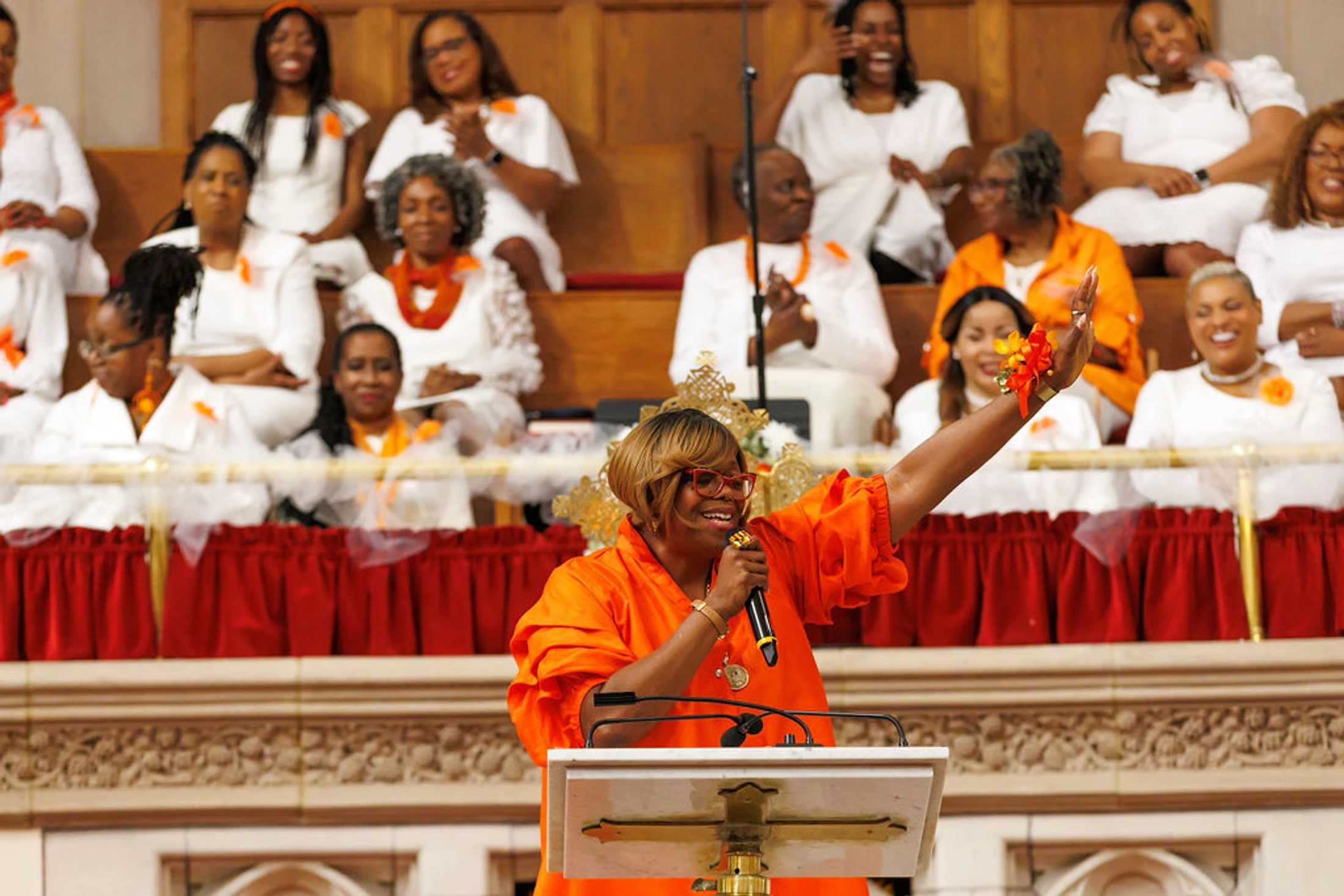 A Black woman in a vibrant orange dress preaches from a pulpit with one arm raised. In the background, a choir dressed in white and orange watches her.
