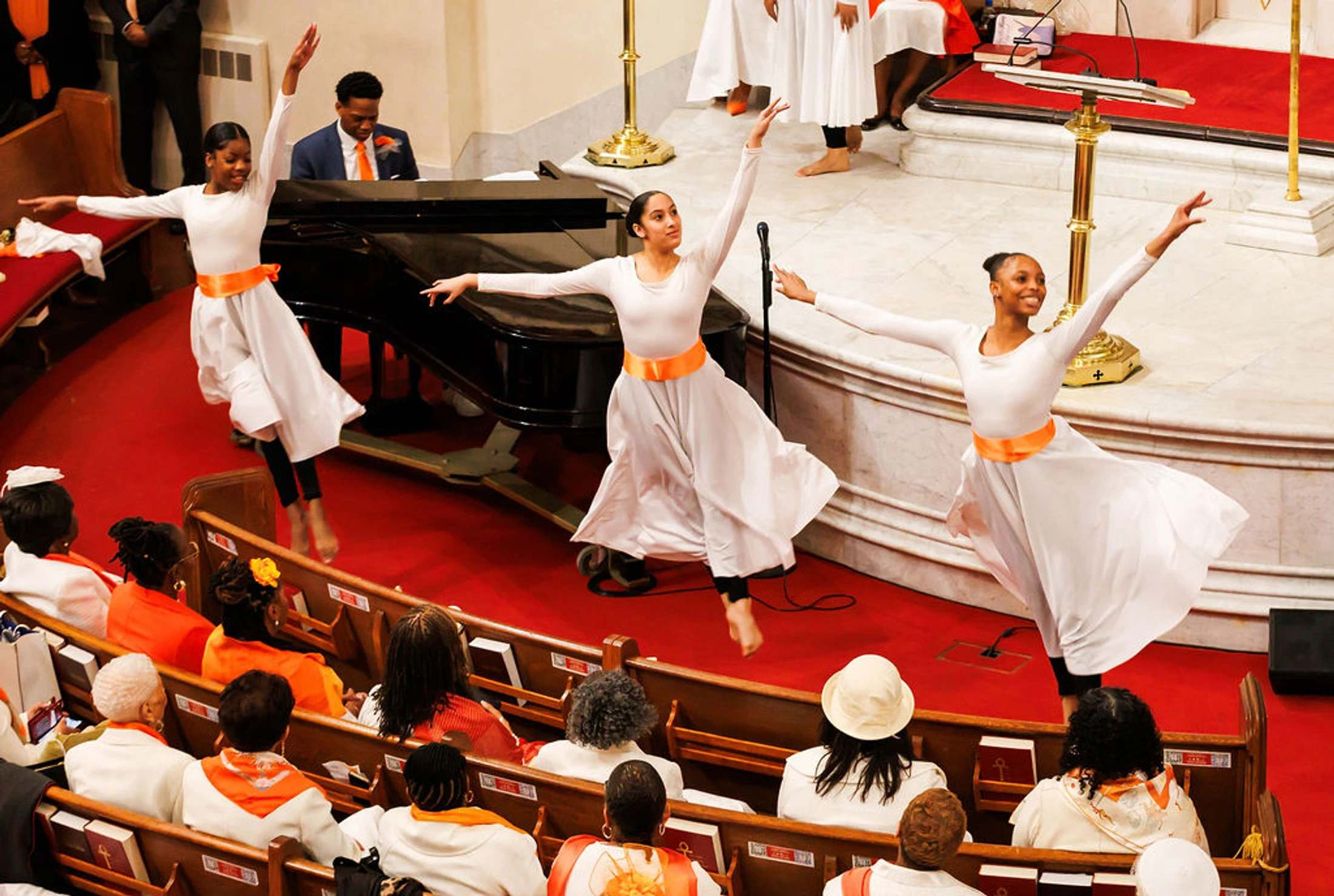 Three young women in matching white and orange outfits perform a graceful dance on a red carpet in a church, as the congregation watches from the pews.