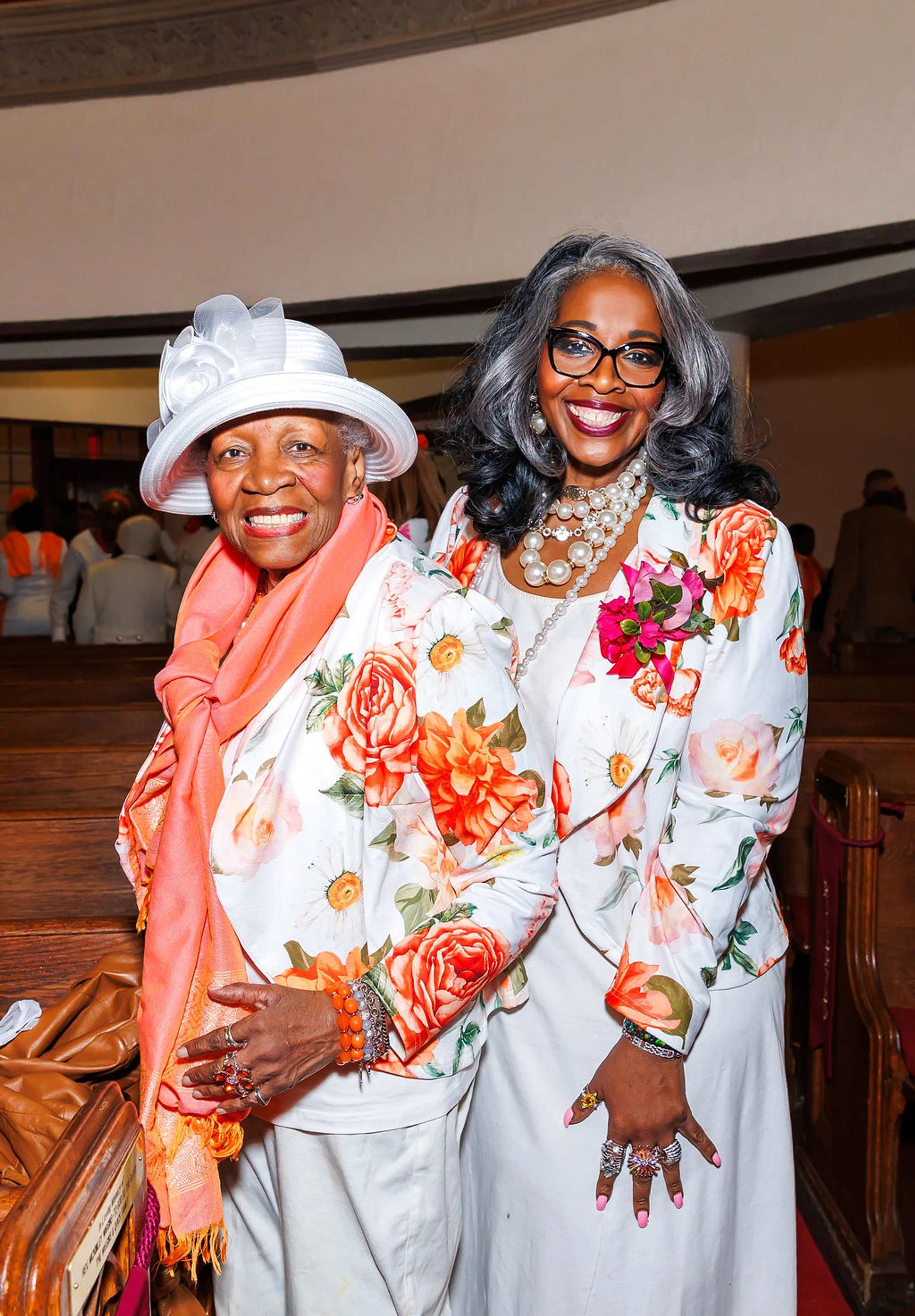 Two elegantly dressed Black women, possibly mother and daughter, smile in a church pew while wearing matching white outfits and vibrant floral jackets.