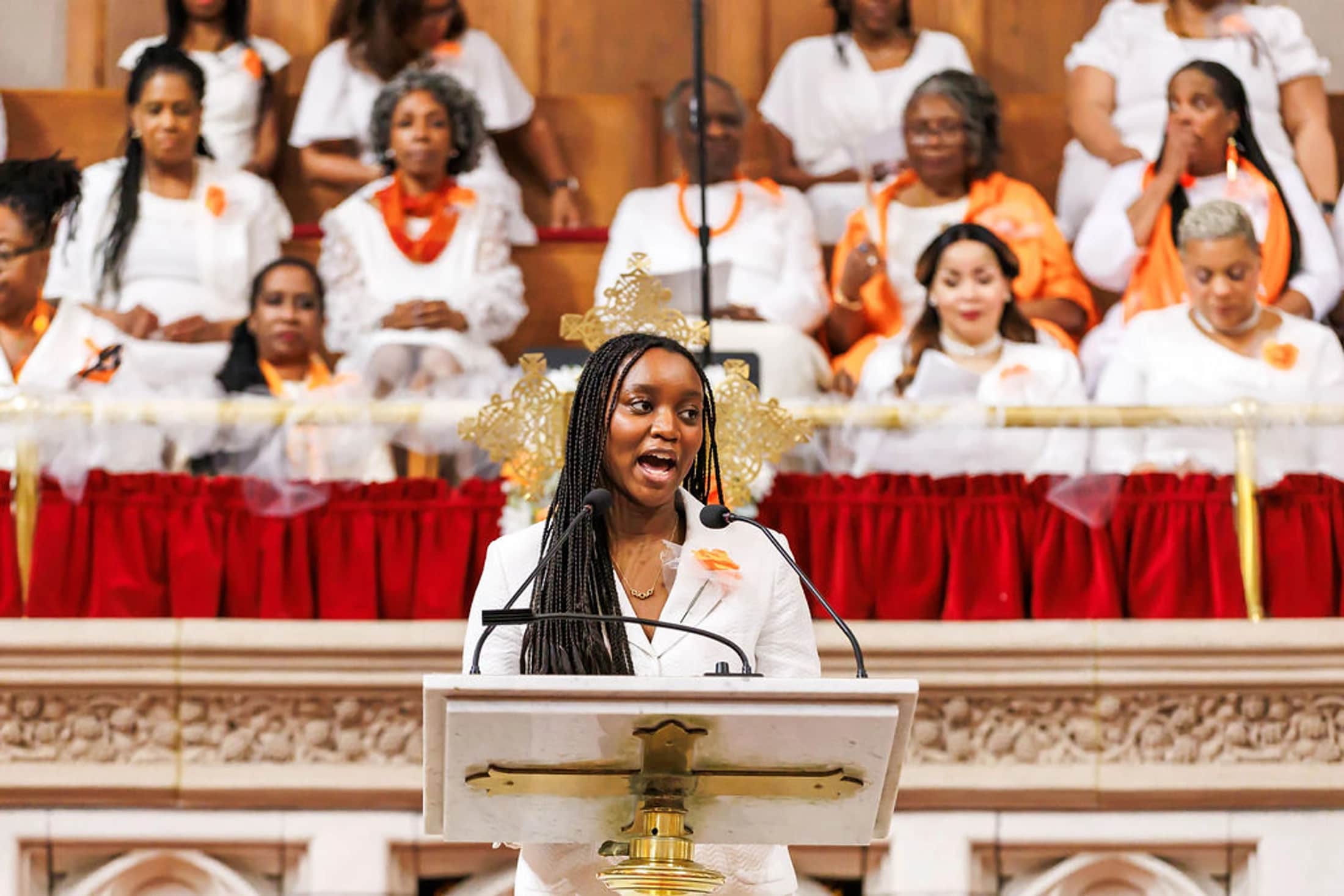 A young Black woman with long braids, wearing a white suit, speaks into microphones at a church pulpit, with a congregation of women in the background.
