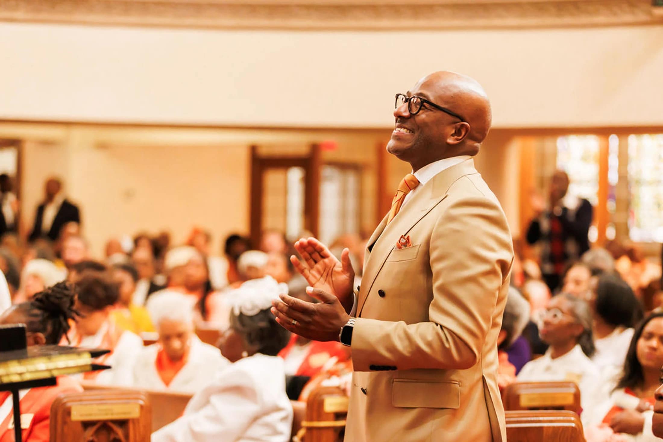 A smiling Black man in a tan suit and glasses stands before a congregation in a church, clapping his hands and looking up joyfully during a service.