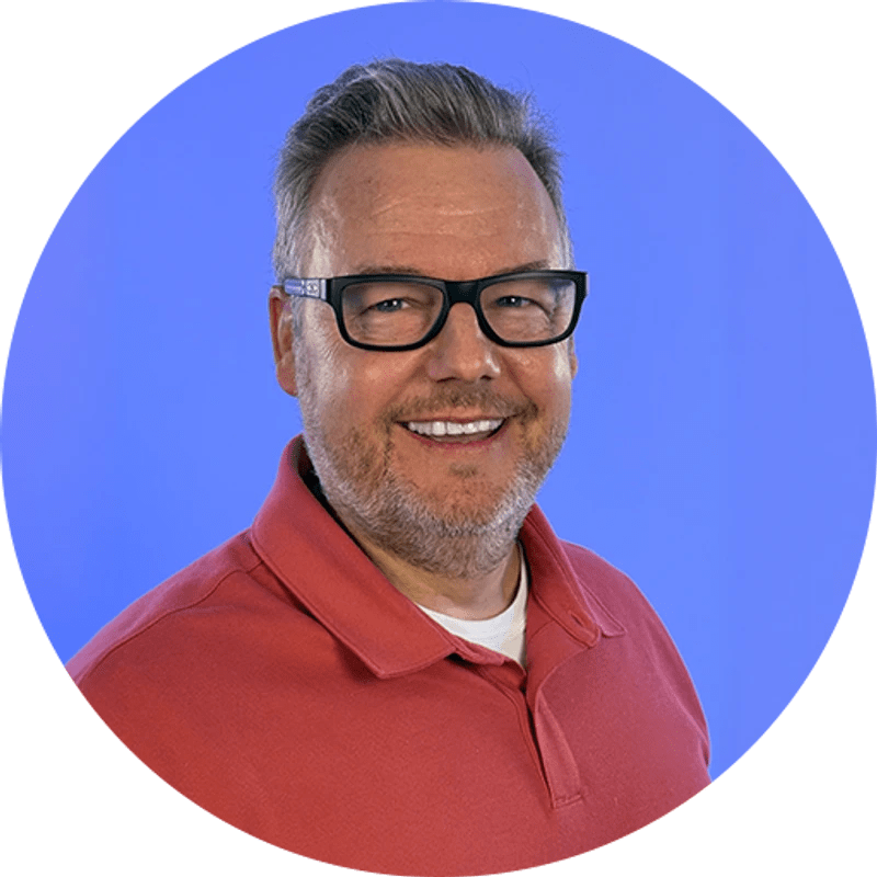 A circular headshot of a smiling, middle-aged man with graying hair and black-rimmed glasses, wearing a red polo shirt against a solid blue background.
