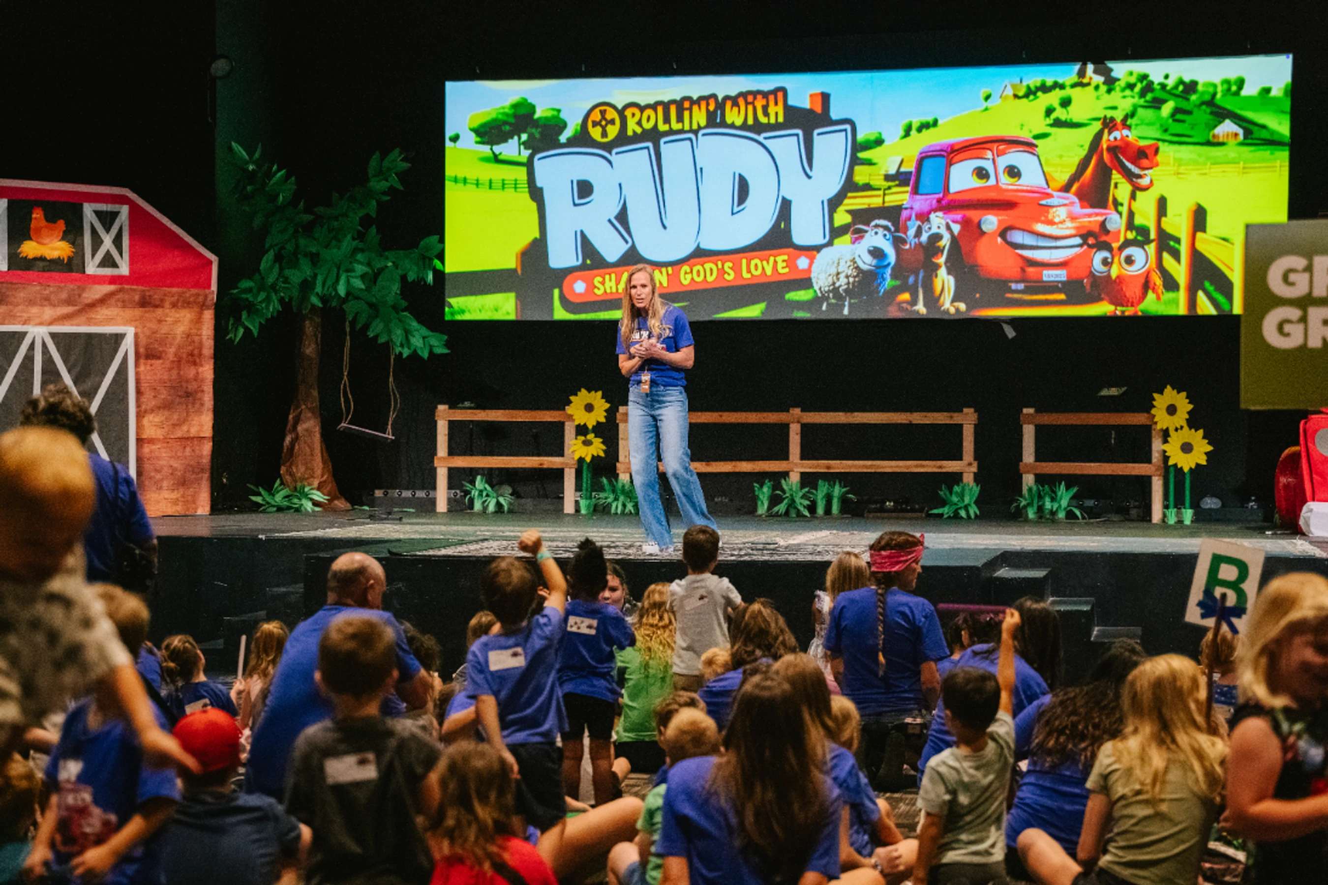 A woman speaks on a farm-themed stage to a large audience of children sitting on the floor. A screen behind her displays a 'Rollin' With Rudy' cartoon.