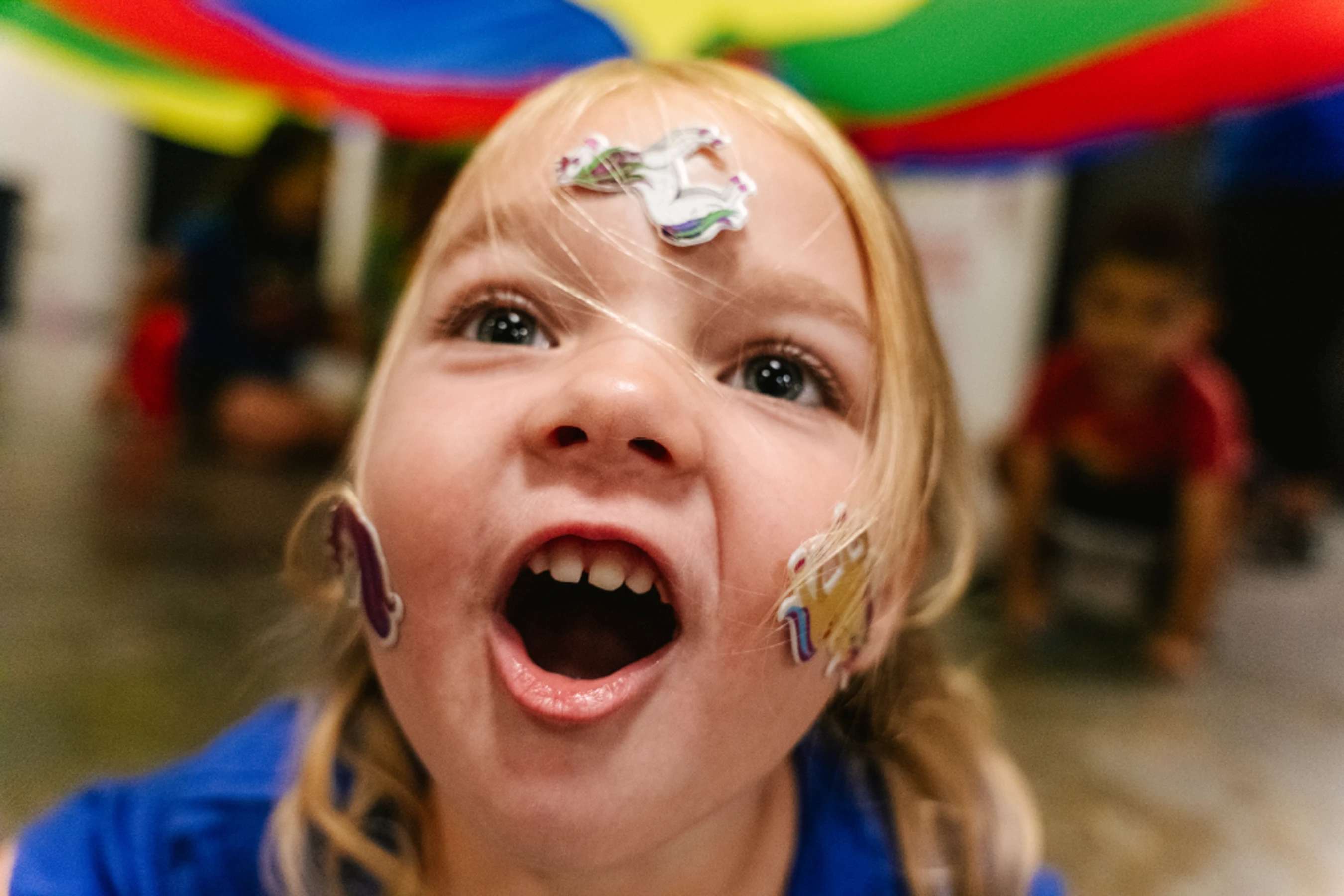 A close-up of an excited young girl with unicorn stickers on her face, looking up with her mouth open while playing under a colorful parachute.