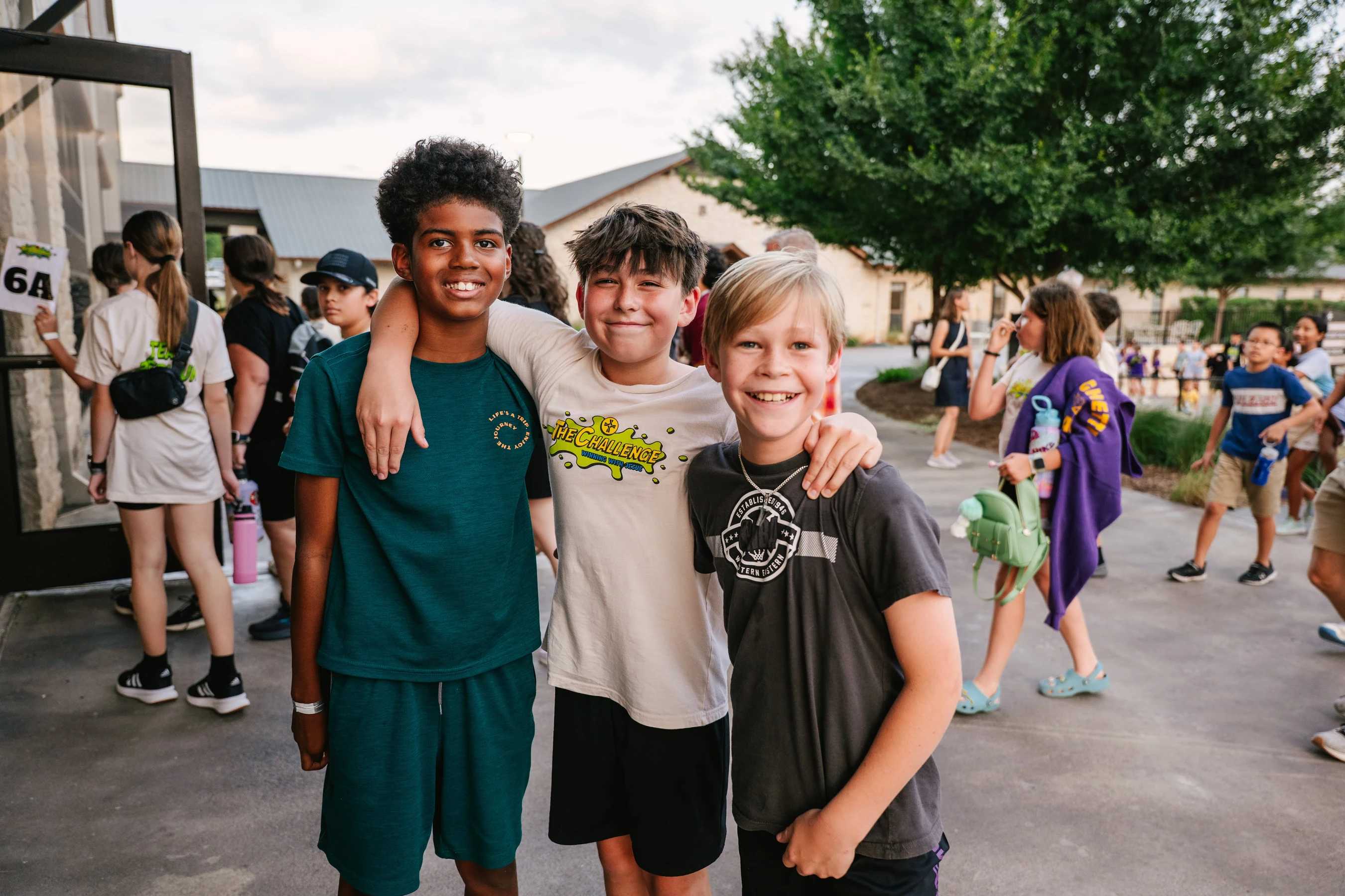 Three diverse young boys stand with their arms around each other, smiling for the camera outside during what appears to be a camp or youth event.