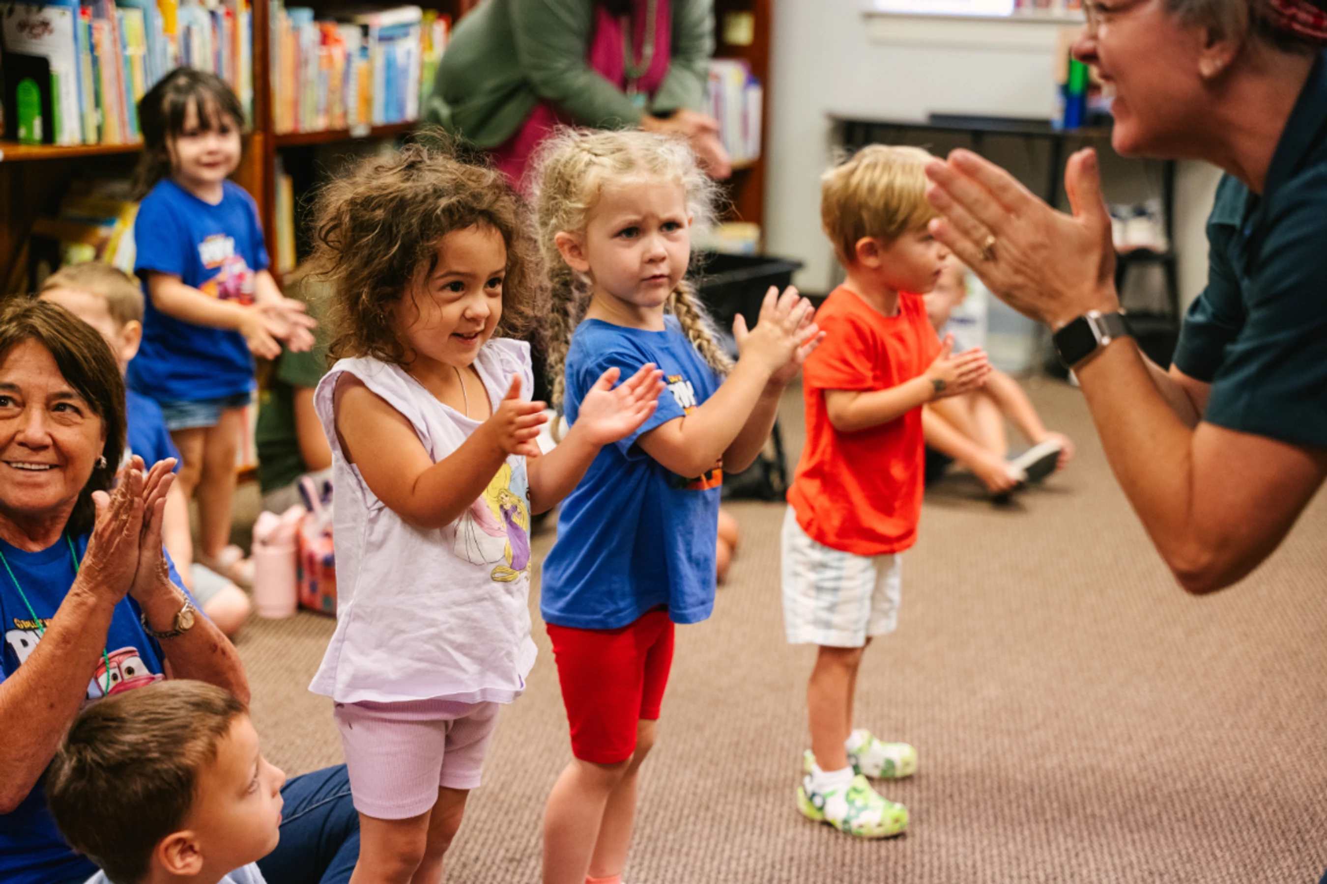 A group of diverse preschoolers stands and claps along with their teachers during a group activity in a classroom with bookshelves in the background.