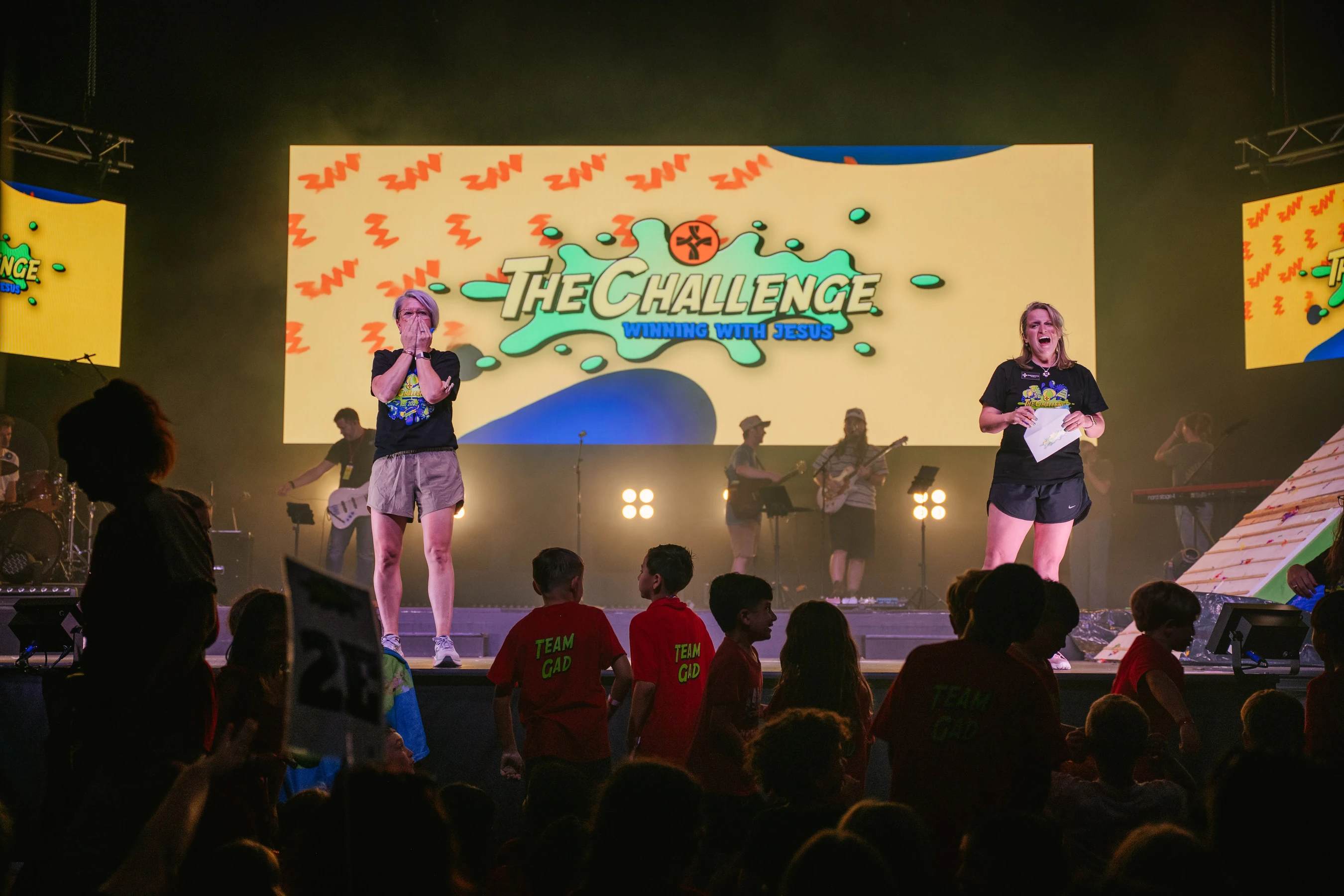 Two women lead a children's event on a stage with a live band. A screen behind them reads 'The Challenge: Winning with Jesus' as kids watch from the audience.