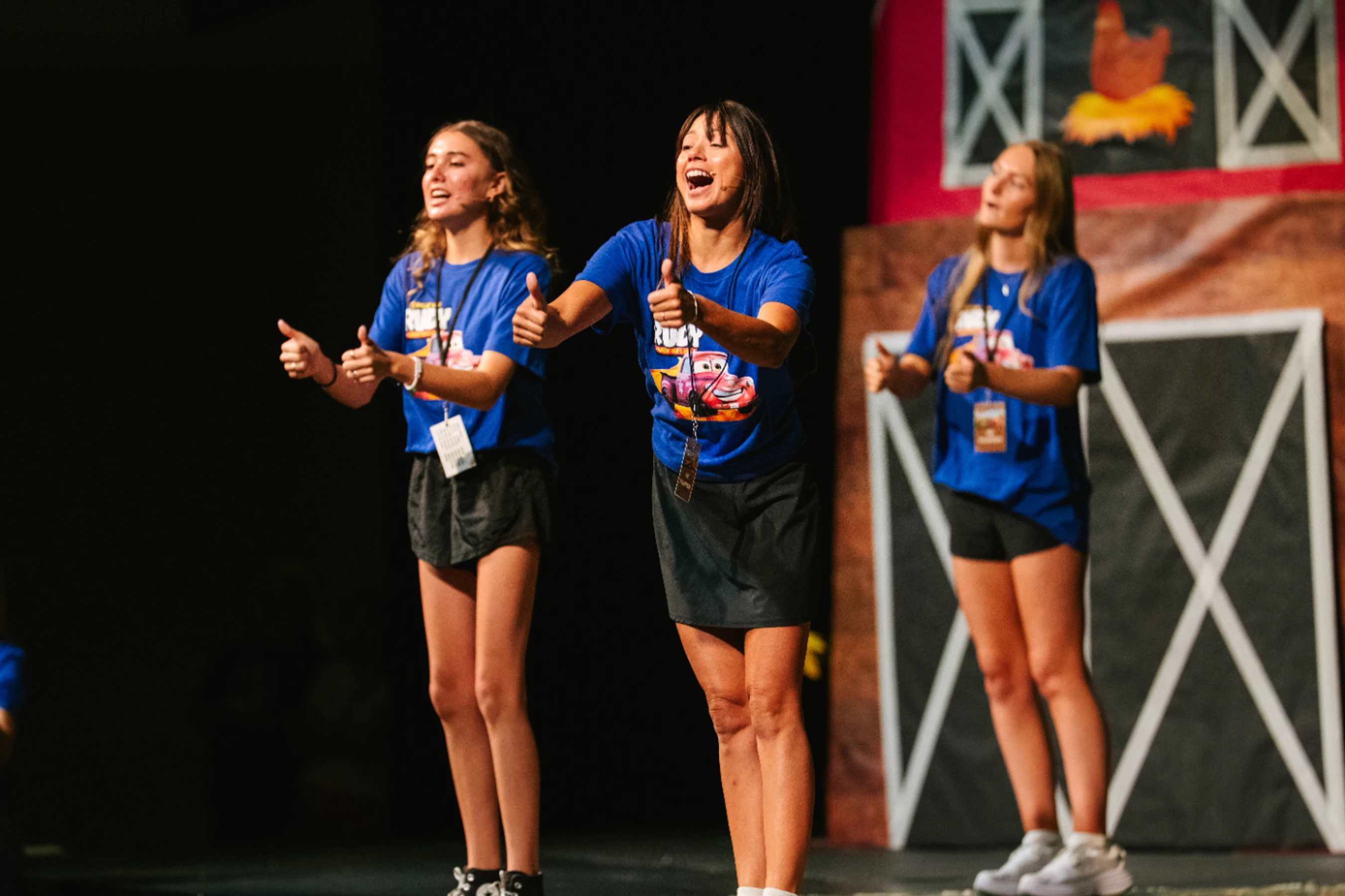 Three young women in matching blue t-shirts stand on a stage, enthusiastically singing and giving thumbs-up gestures towards the audience.