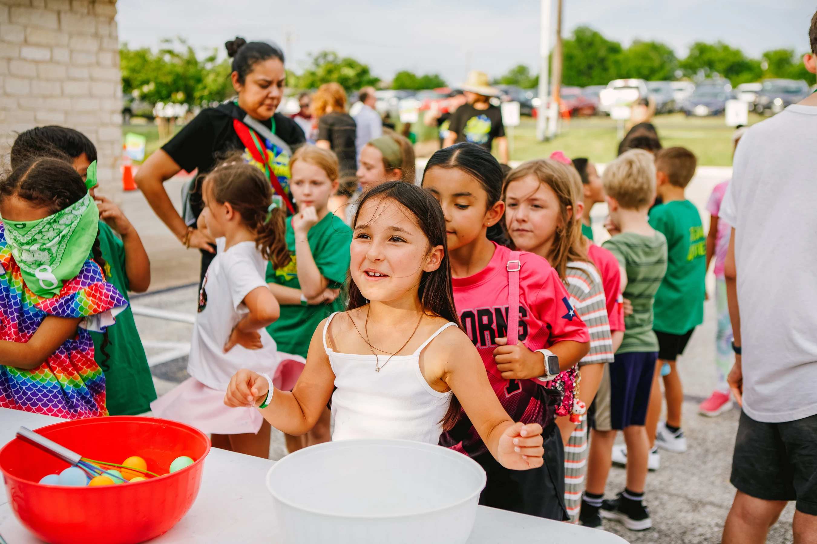 A young girl in a white top smiles happily at the front of a line of children waiting to participate in a game with bowls and colorful balls outdoors.