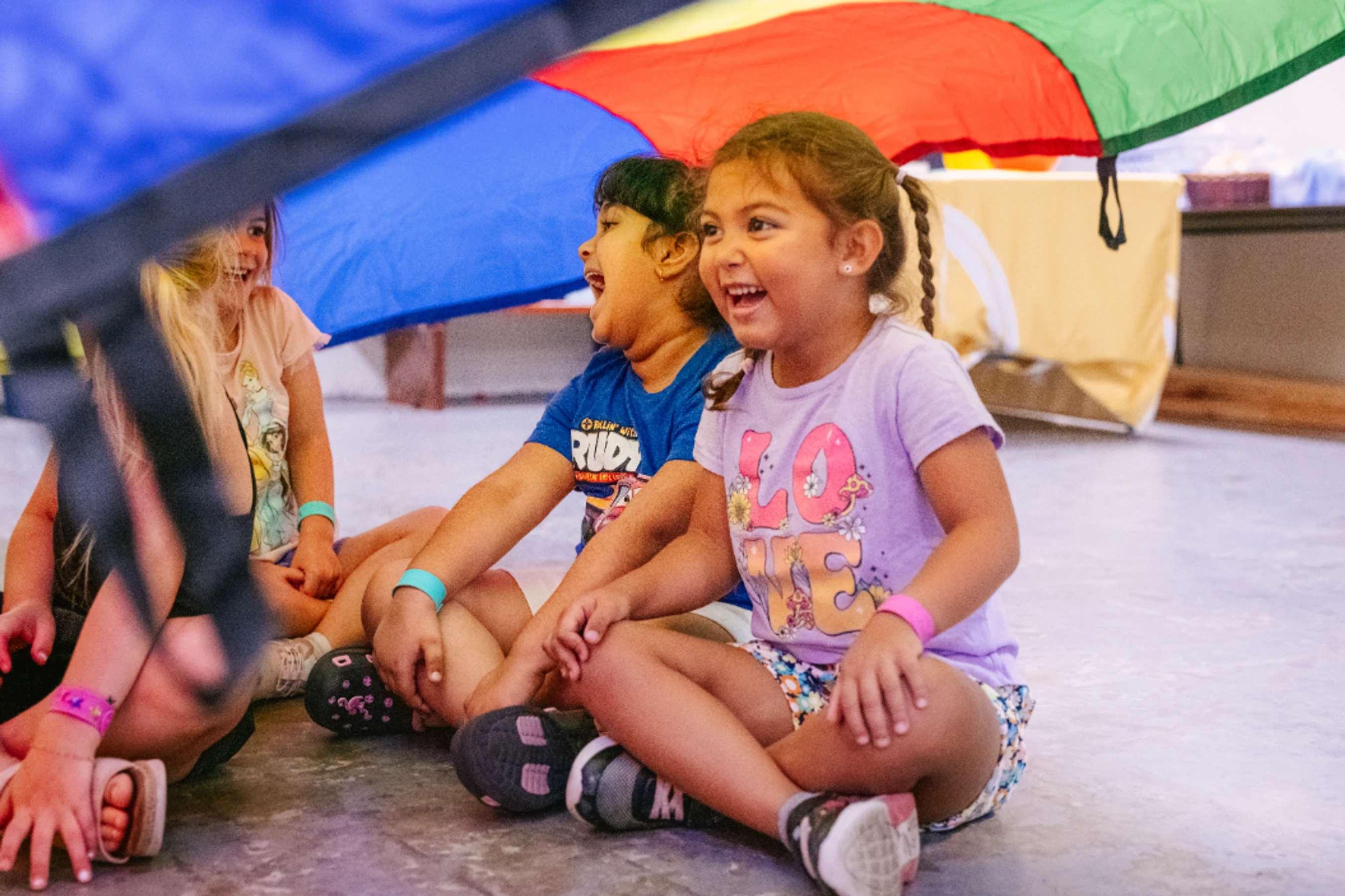 A group of young children sit cross-legged on the floor under a large, colorful rainbow parachute, smiling and laughing together during a fun activity.