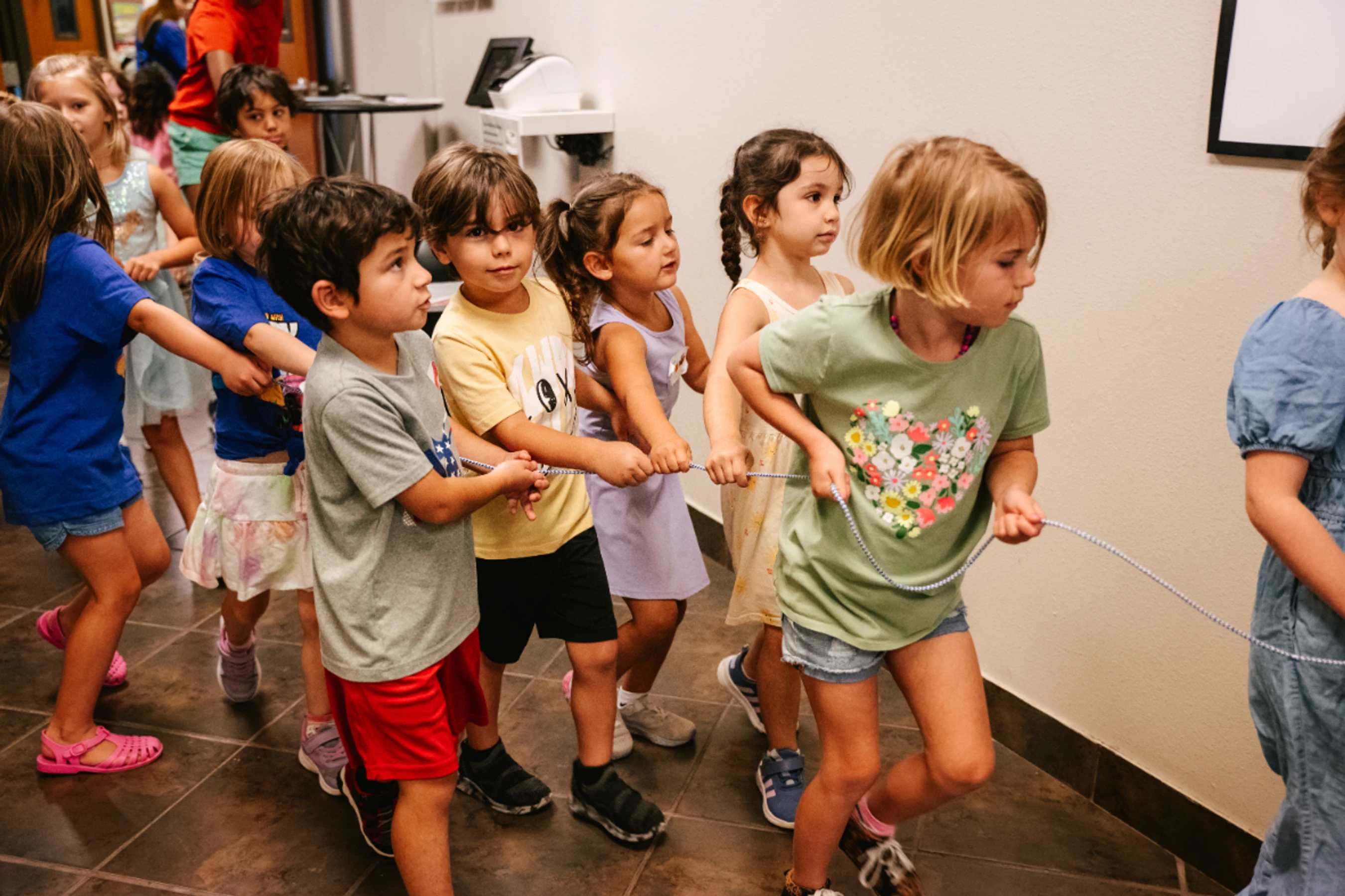 A group of diverse young children walk in a single file line down a hallway, each holding onto a shared patterned rope to stay together.